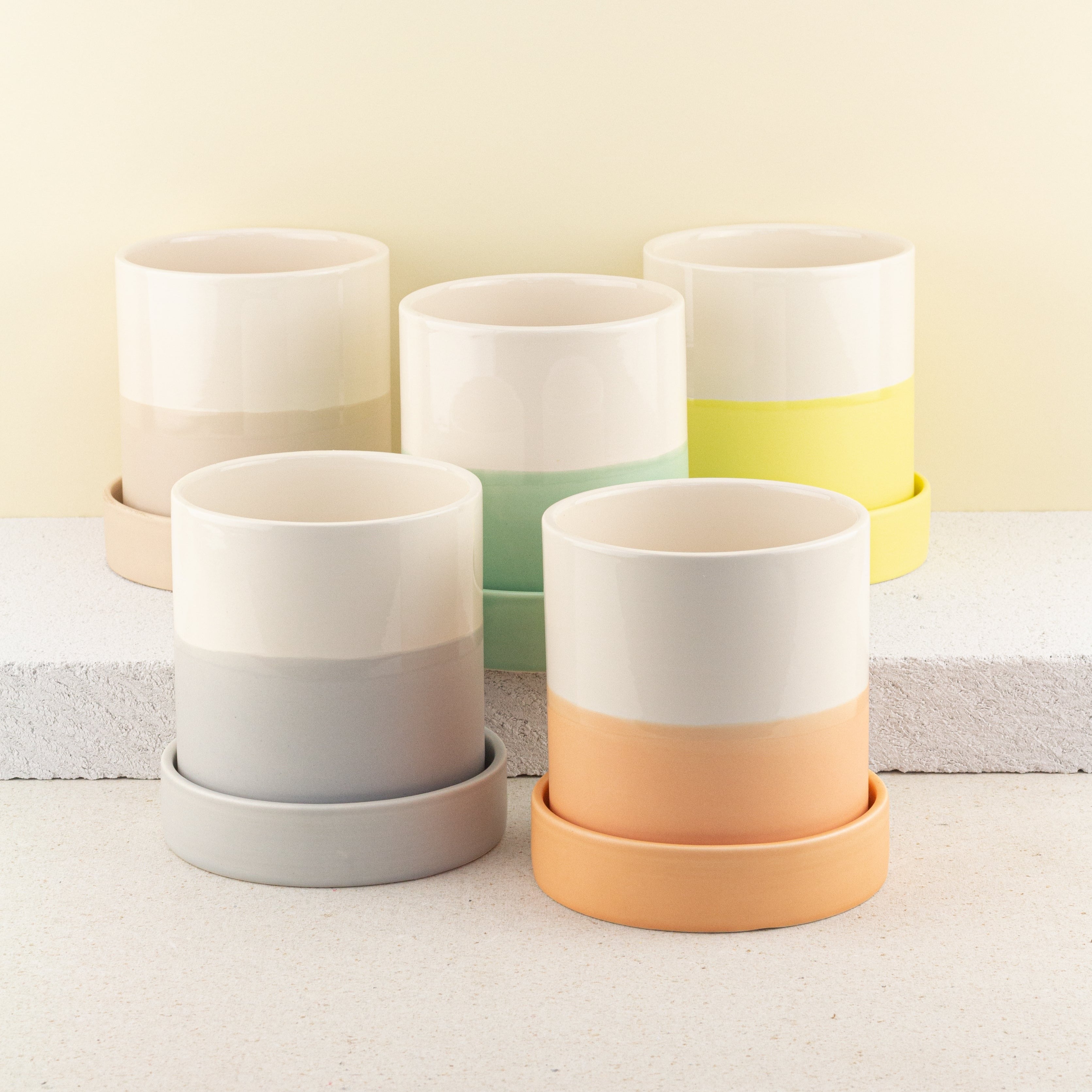 Fünf zweifarbige Pflanzentöpfe in Beige, Grau, Grün, Gelb und Orange auf gestuften, hellen Podesten vor pastellgelbem Hintergrund.