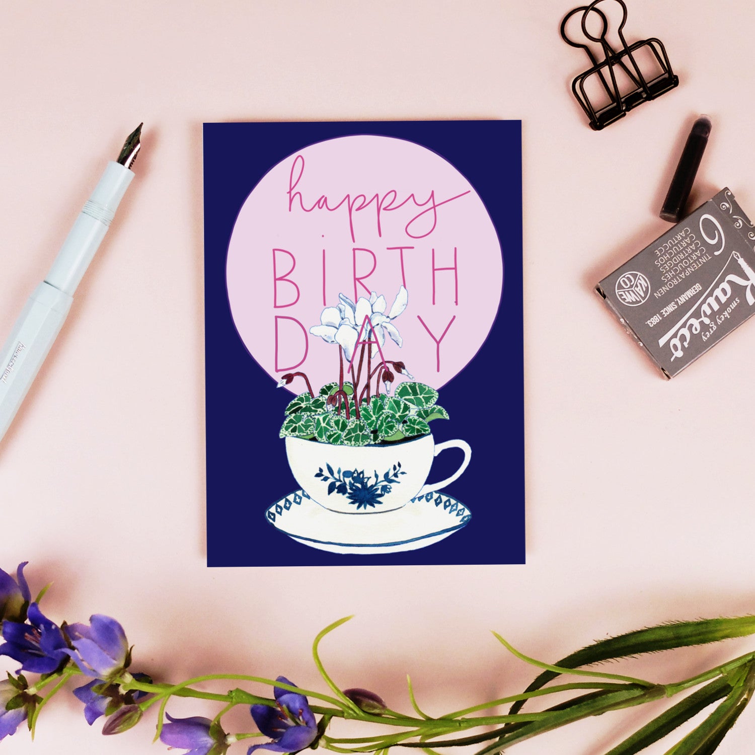 Eine Grußkarte mit der Aufschrift "Happy Birthday" über einer Teetasse mit weißen Blumen und grünen Blättern, die auf einer rosafarbenen Fläche mit einem Stift, einer Büroklammer, einem Radiergummi und lila Blumen in der Nähe steht.