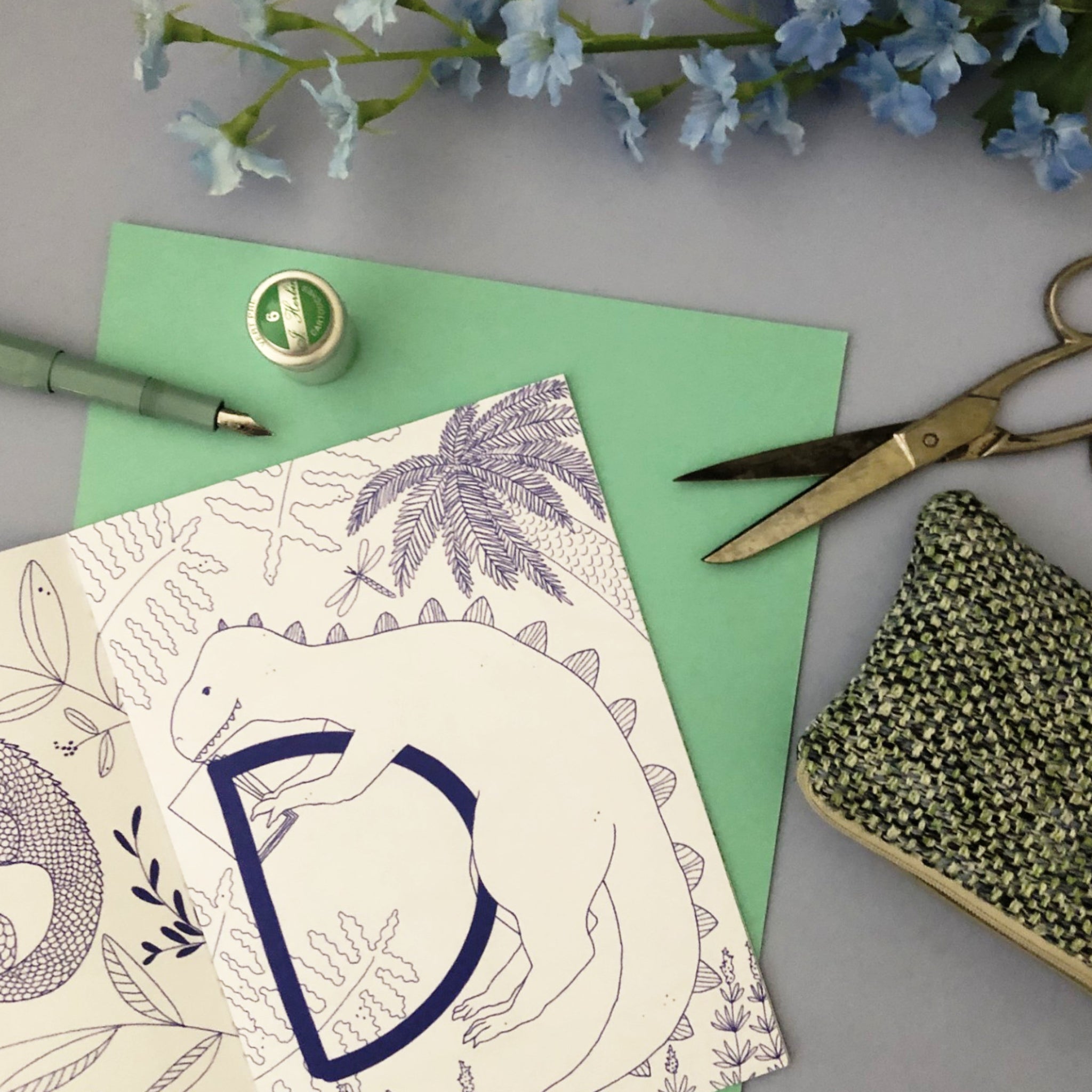 Karte mit Dinosaurier-Illustration, Stift, Schere, Fadenrolle und blauen Blumen auf hellgrauem Hintergrund.