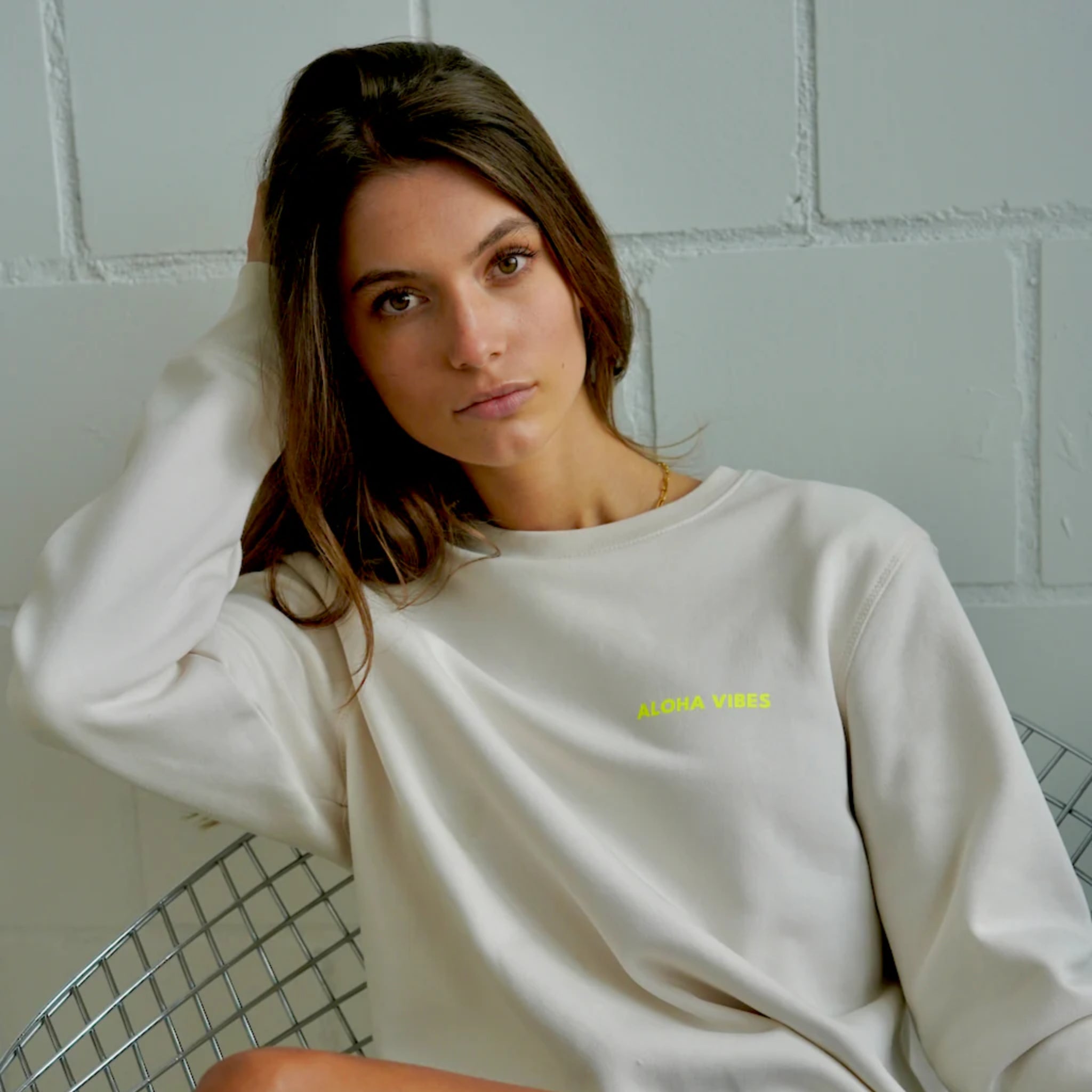 Eine Frau mit langen braunen Haaren und einem cremefarbenen Sweatshirt, auf dem in gelben Buchstaben ALOHA VIBES steht, sitzt auf einem Metallstuhl und schaut in die Kamera. Der Hintergrund ist eine hellgrau gestrichene Ziegelwand.