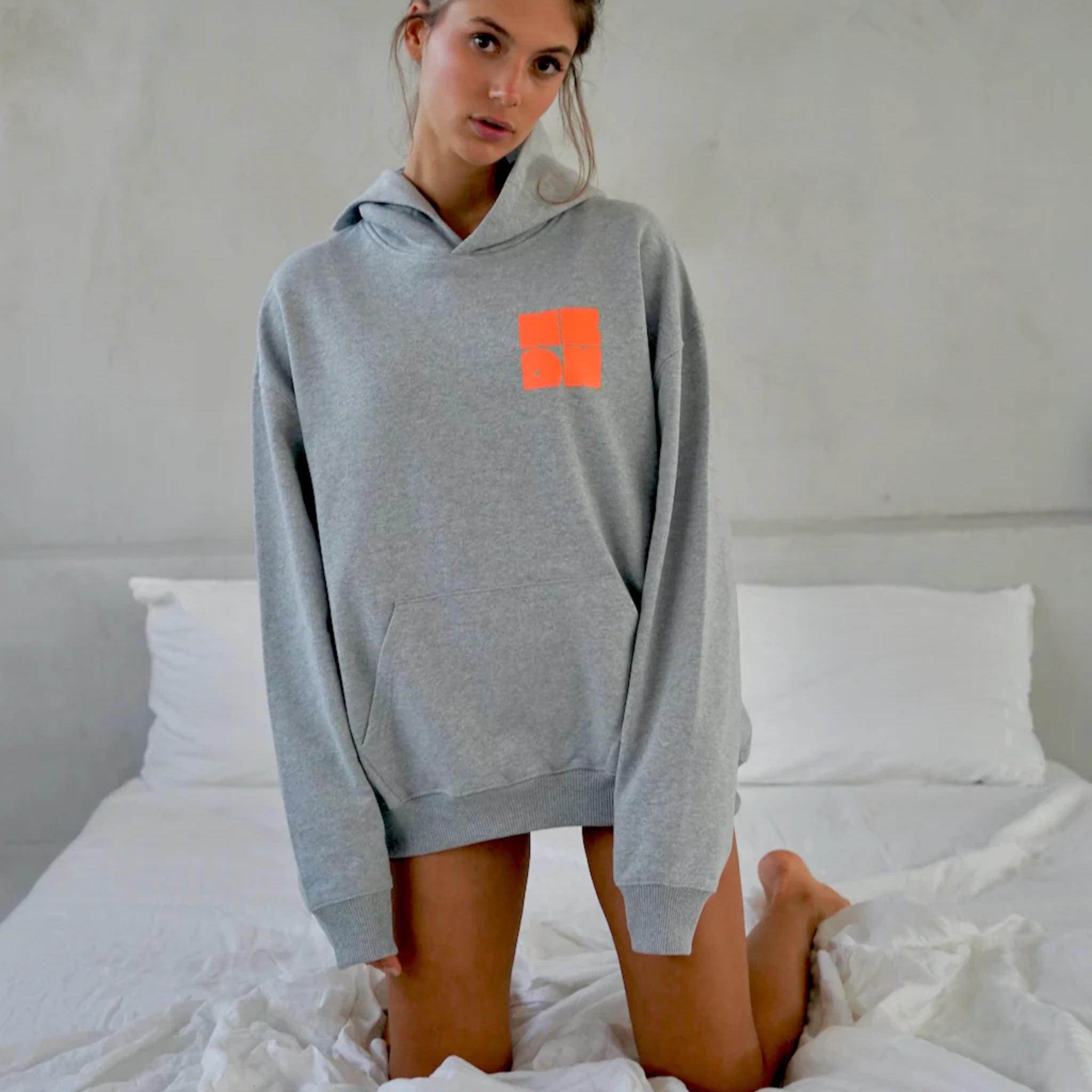 Eine junge Frau kniet auf einem Bett und trägt einen übergroßen grauen Kapuzenpullover mit einem kleinen orangefarbenen quadratischen Muster auf der Brust vor einem schlichten hellen Wandhintergrund.