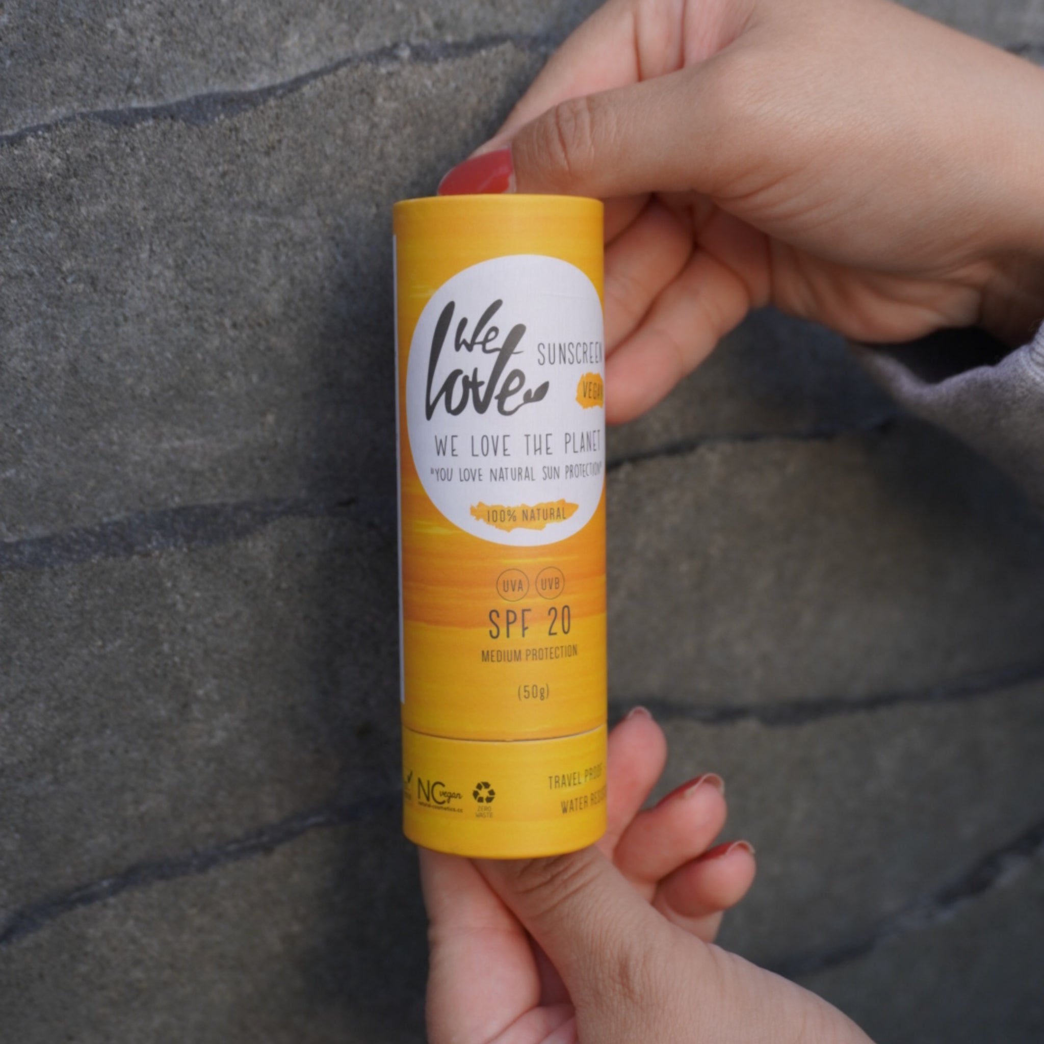 Eine Hand hält eine gelbe Sonnencreme mit der Aufschrift "We Love the Planet" und SPF 20 vor einer grauen Wand.