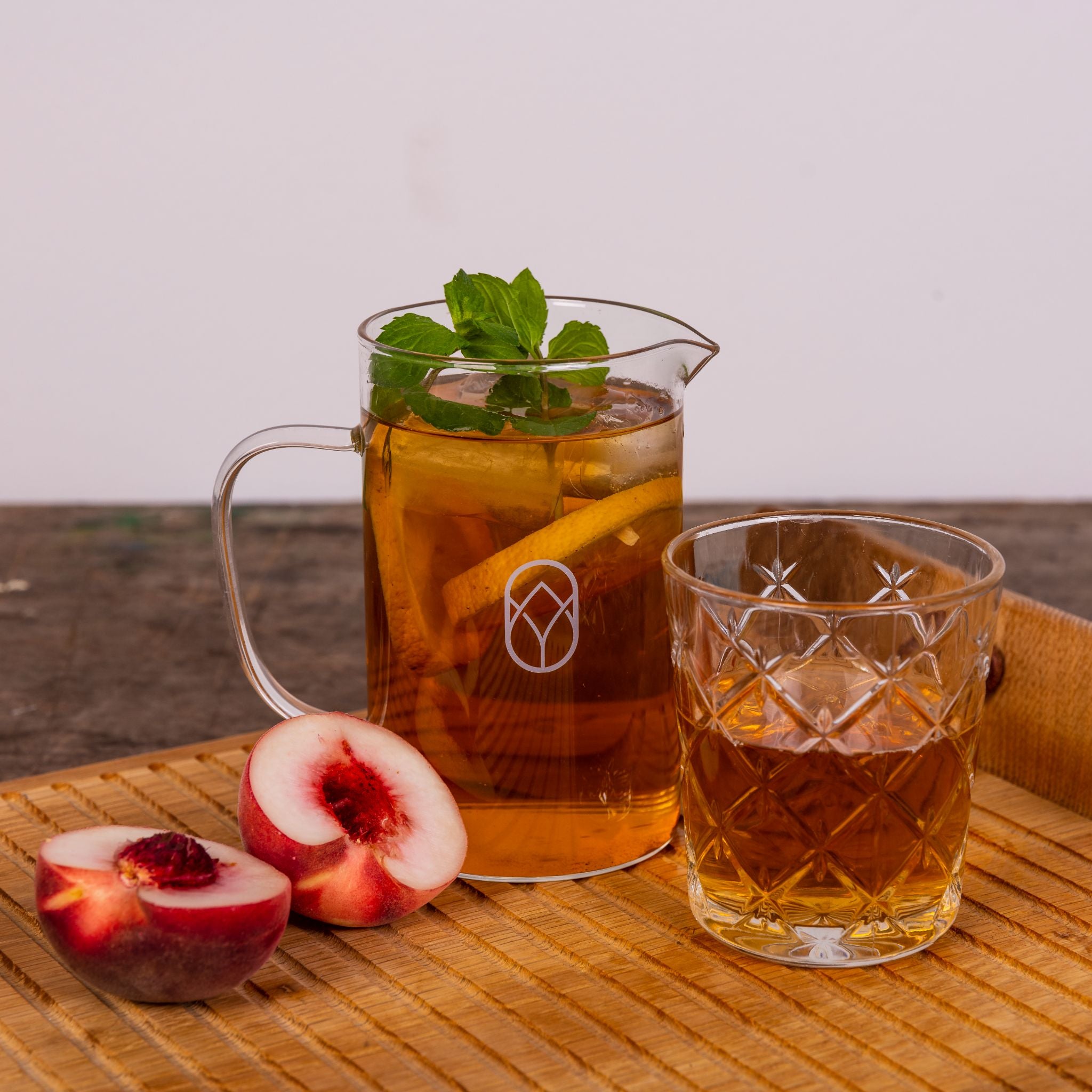 Glaskrug und Glas mit Eistee auf Holztablett neben halben Pfirsichen, Krug mit Zitronenscheiben und Minze garniert.