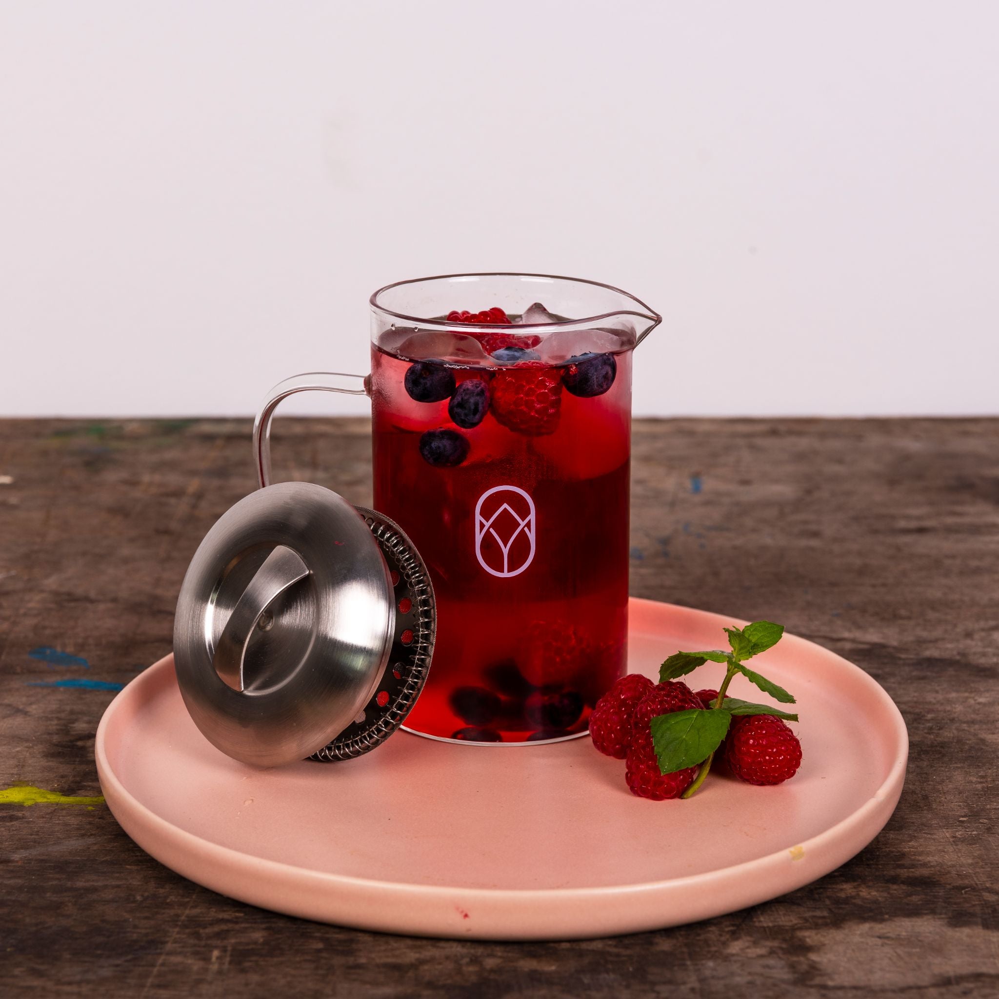 Ein Glaskrug mit roten und blauen Beeren, daneben ein Deckel und ein Teller mit Himbeeren und Minzblättern.