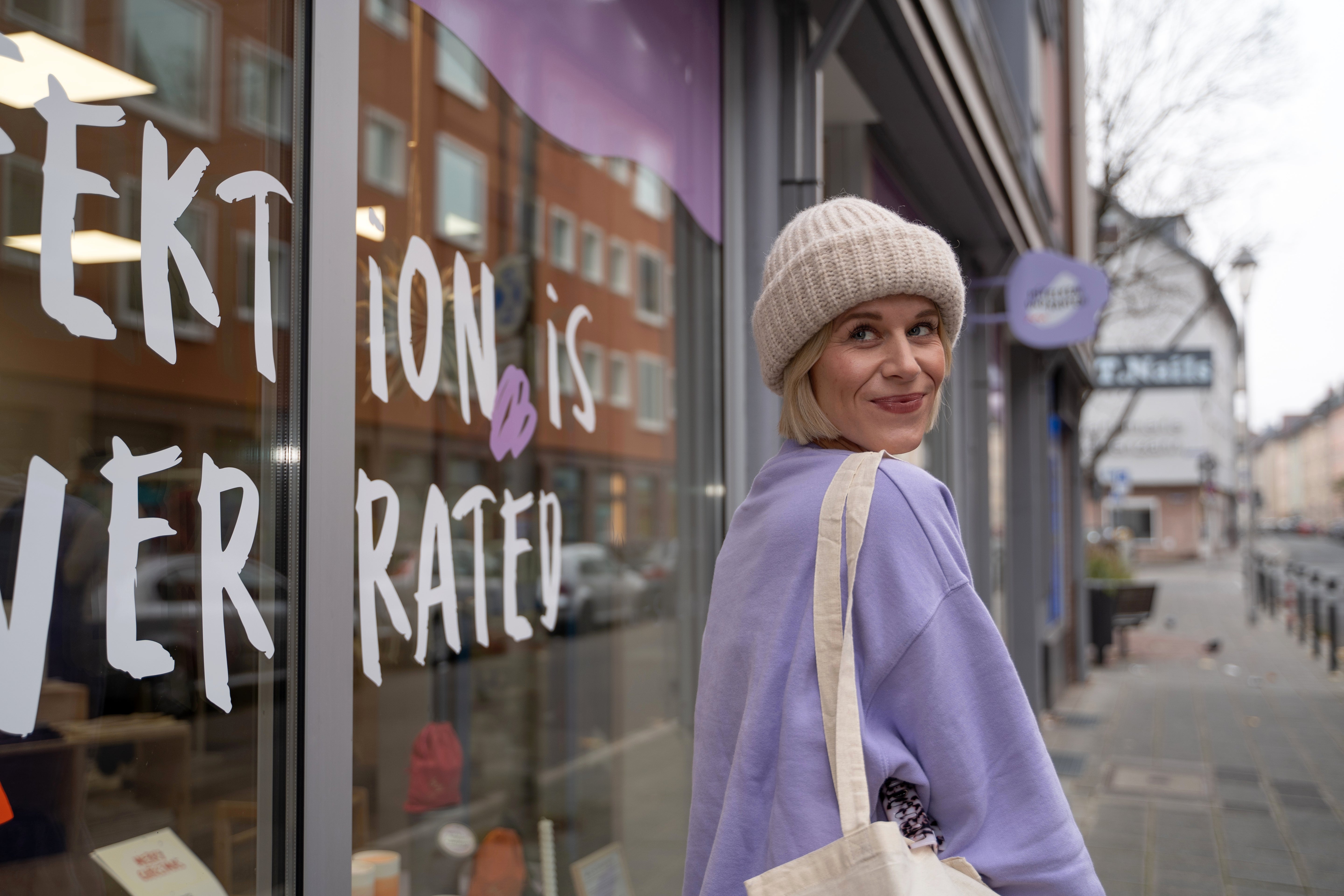 Frau in lila Pullover und Mütze steht vor einem Schaufenster mit dem Satz "Perfection is overrated", lächelnd zur Kamera.
