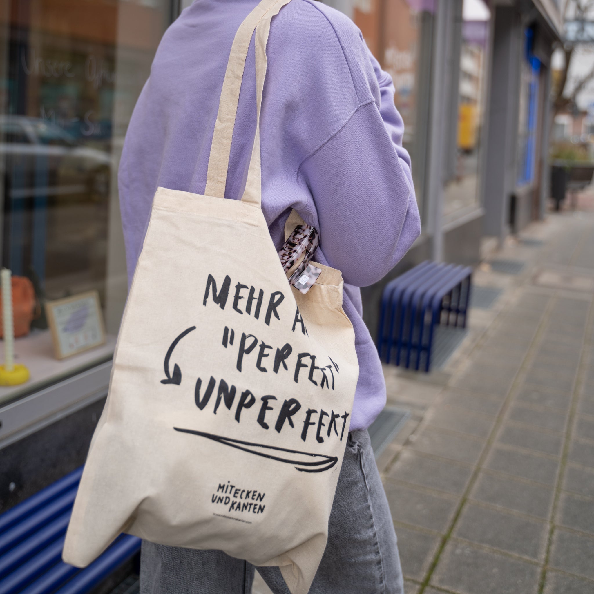 Person mit lila Pullover trägt eine Stofftasche, auf der „Mehr als perfekt unperfekt“ steht, auf der Straße.
