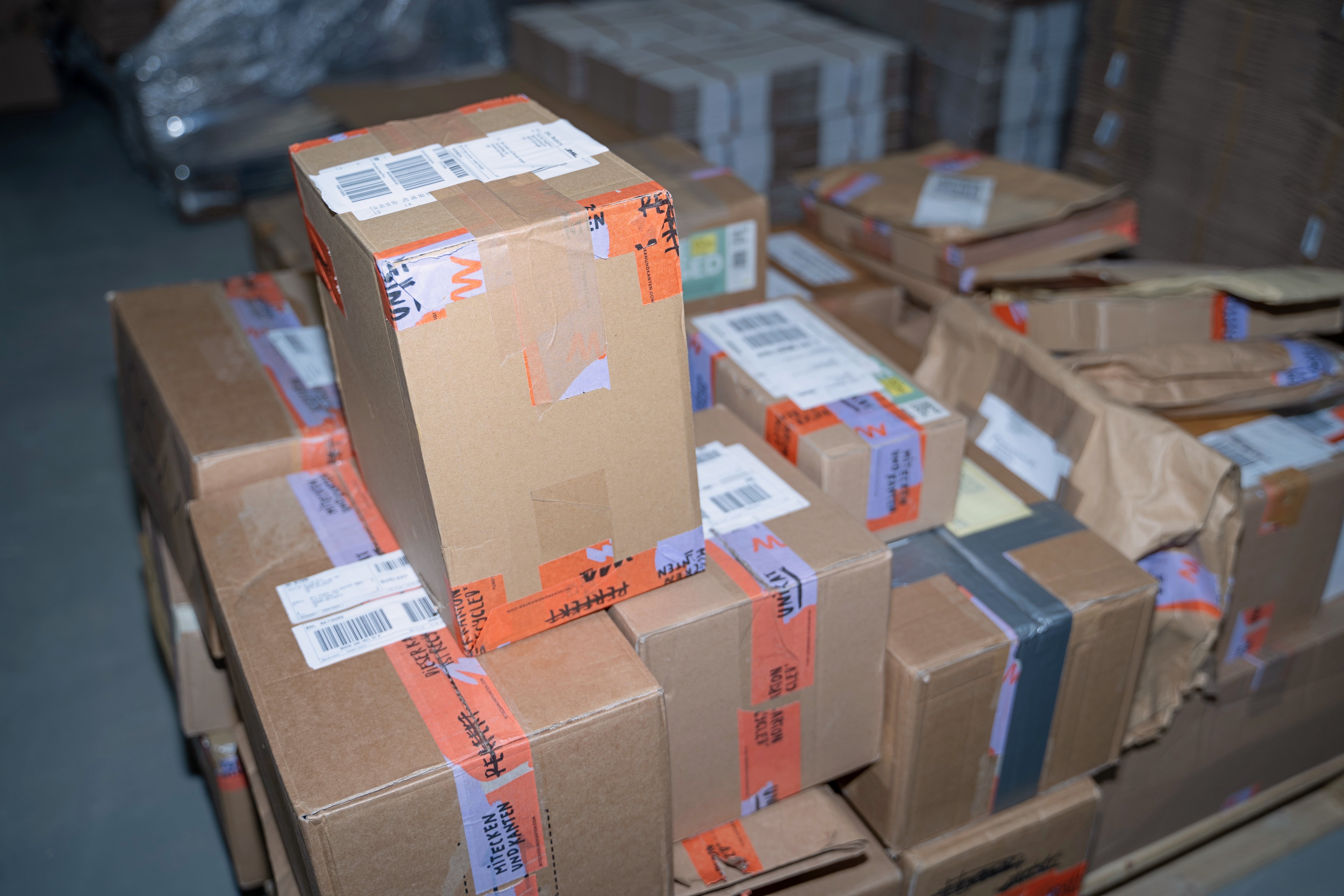 Ein Stapel von Paketen in einem Lager mit Versandetiketten und orangefarbenem Klebeband versehen.