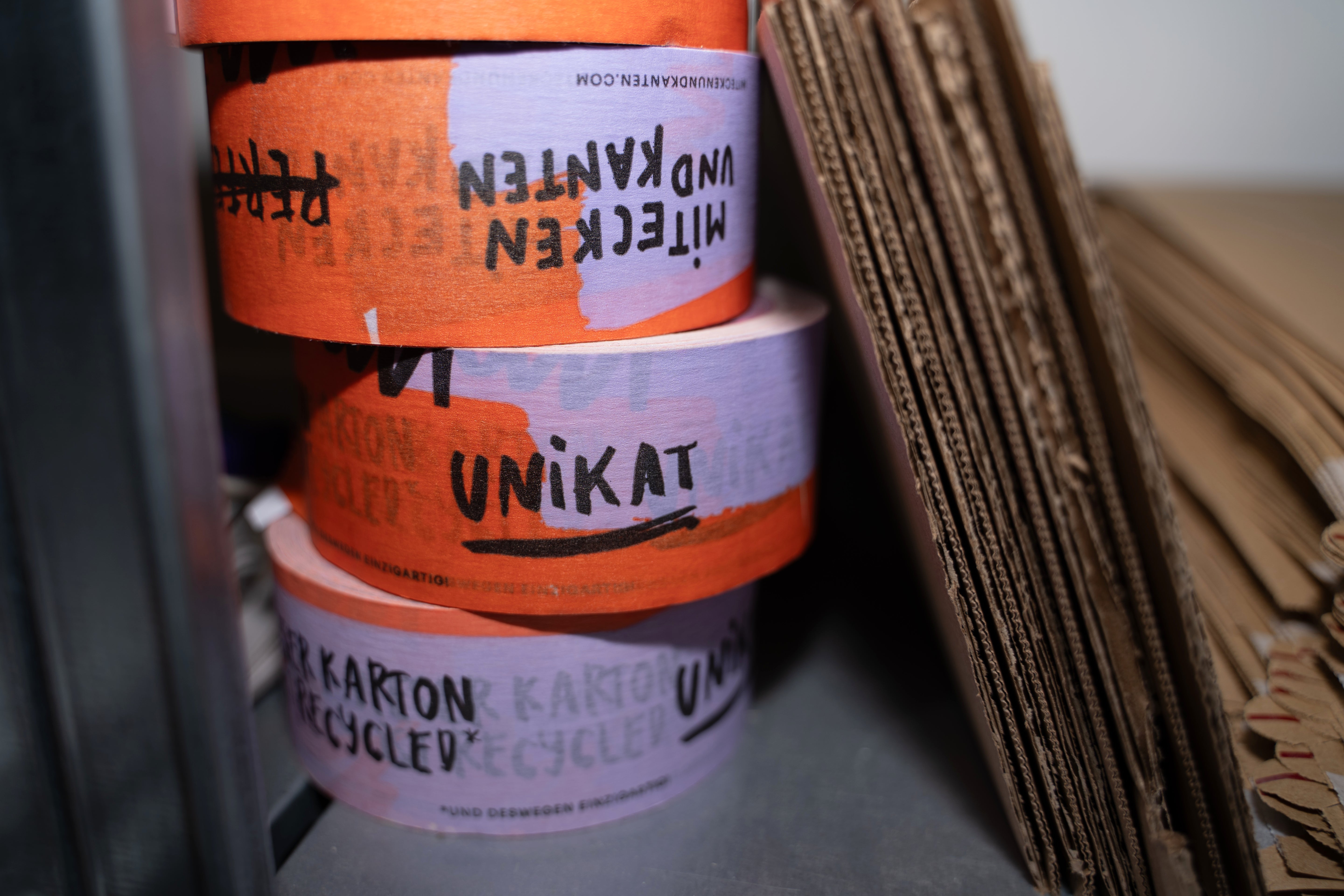 Stapel von buntem Klebeband mit der Aufschrift "Unikat" neben gestapeltem Wellkarton auf einem Regal.