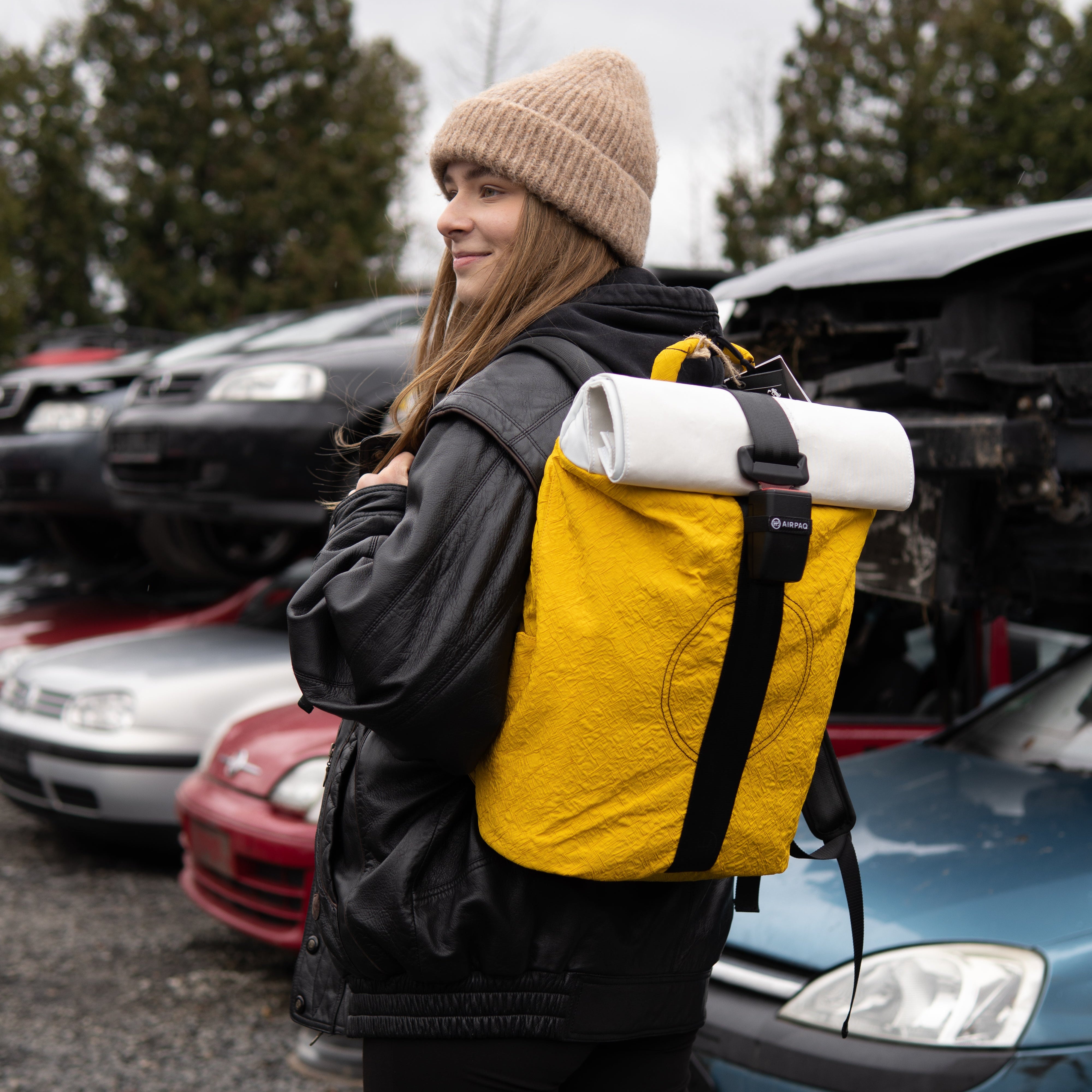 Eine Frau mit einer beigen Strickmütze, einer schwarzen Jacke und einem leuchtend gelben Rucksack steht im Freien auf einem Schrottplatz mit gestapelten, beschädigten Autos im Hintergrund. Sie lächelt und blickt zur Seite.