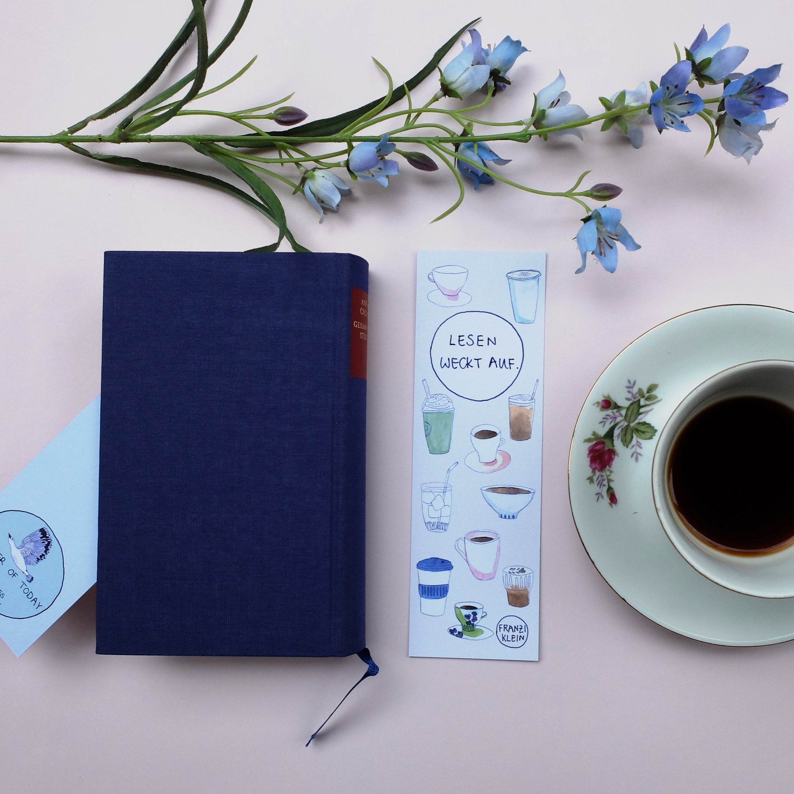 Ein blaues Buch, zwei illustrierte Lesezeichen, eine weiße Tasse mit schwarzem Kaffee auf einer geblümten Untertasse und blaue Blumen sind auf einer hellen Fläche angeordnet.