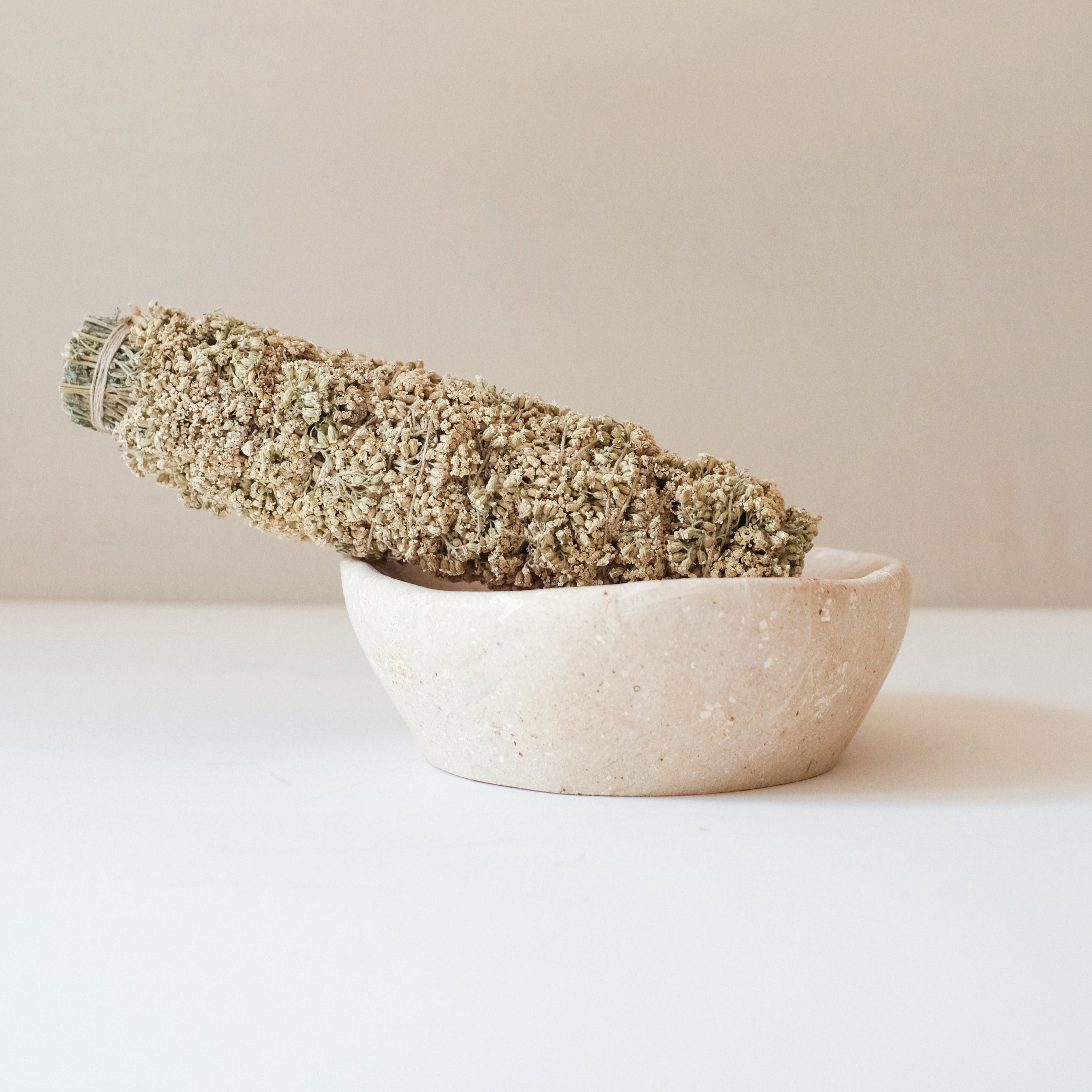 Getrocknetes Bündel aus Kräutern oder Blumen liegt schräg in einer kleinen, flachen Schale aus hellem Stein.