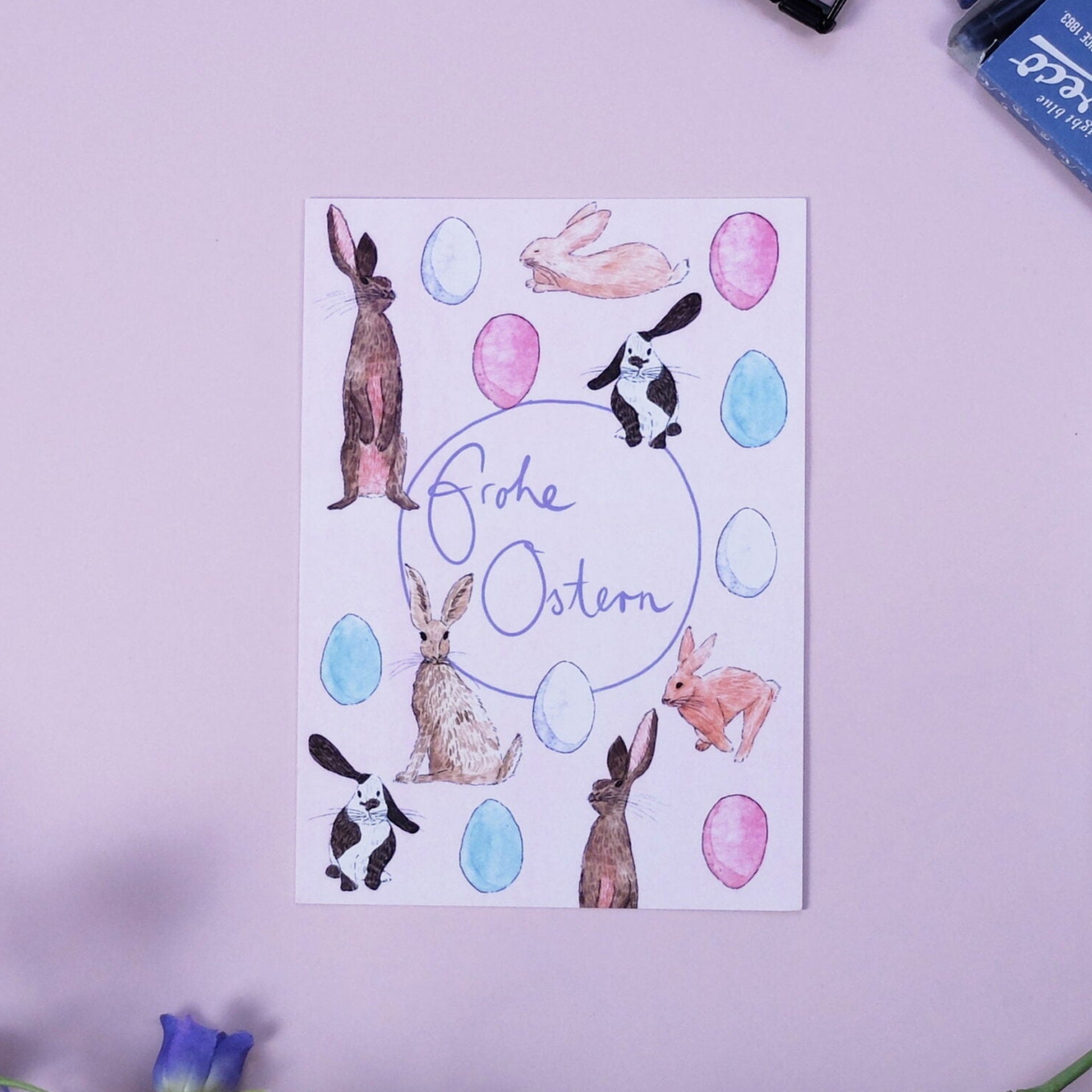 Eine Grußkarte mit "Frohe Ostern", umgeben von aquarellierten Hasen und Eiern, auf einer hellrosa Fläche mit einem blauen Stift, einem Tintenfass, einer schwarzen Büroklammer und lila Blumen in der Nähe.