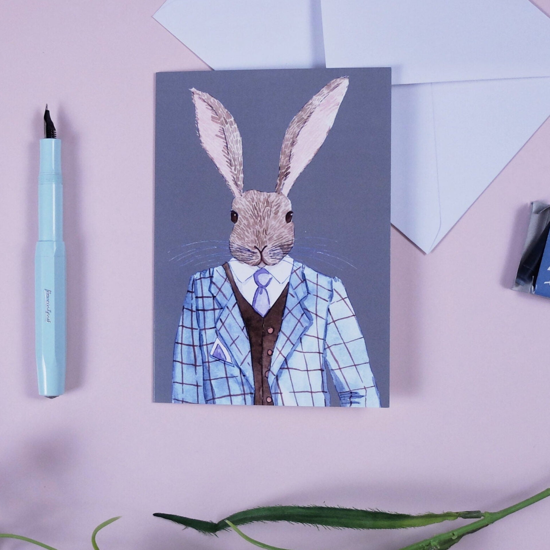 Eine Grußkarte mit der Illustration eines Kaninchens in einem blaukarierten Anzug mit Krawatte liegt auf einer rosa Fläche, umgeben von einem Umschlag, einem hellblauen Füllfederhalter und einer grünen Pflanze.
