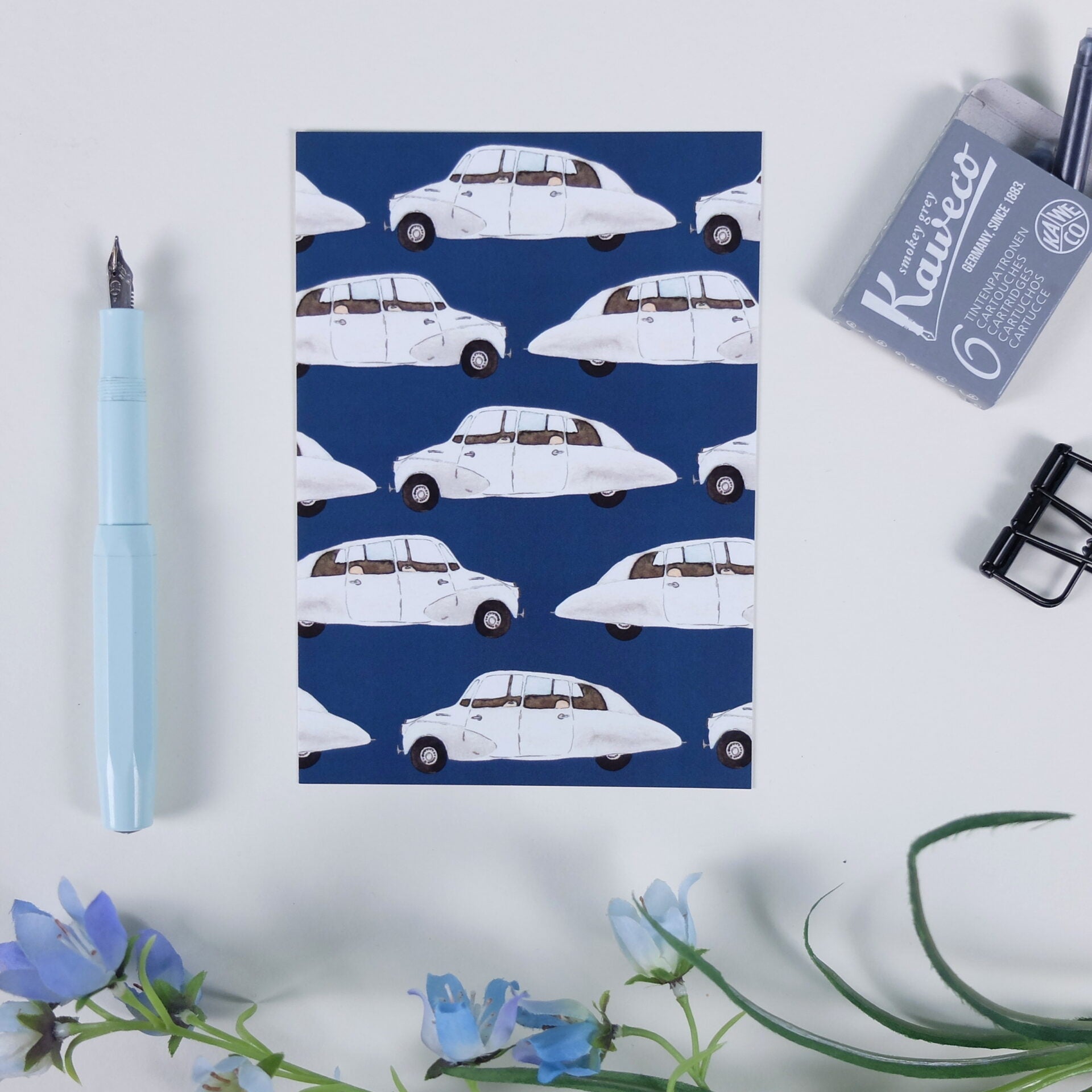 Papier mit Muster aus weißen Oldtimer-Autos, daneben ein türkisfarbener Füller und Büroklammern auf einem Tisch.