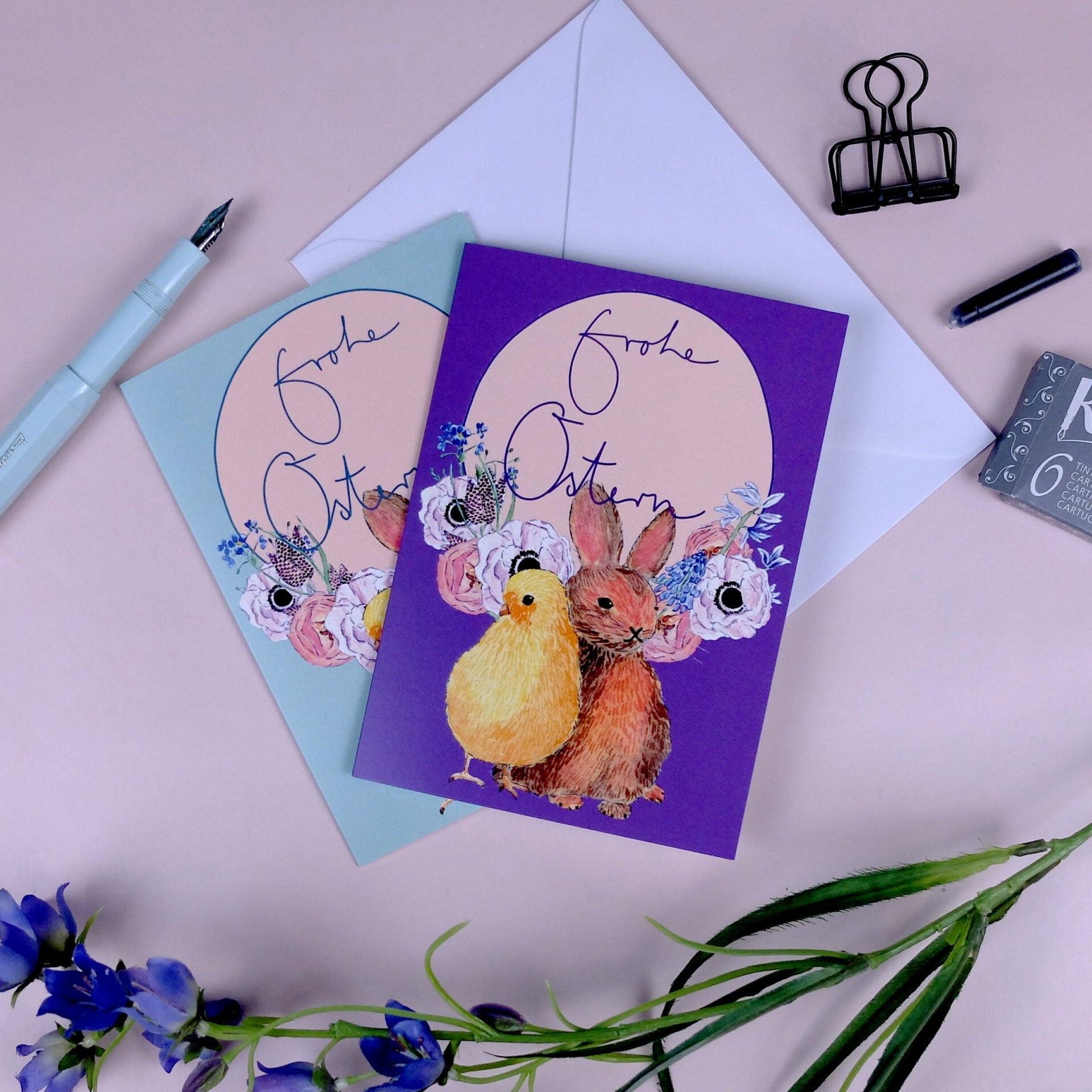 Zwei illustrierte Osterkarten mit dem deutschen Text "Frohe Ostern" zeigen ein Küken und einen Hasen mit Blumen. Die Karten befinden sich auf einer rosafarbenen Fläche, neben einem Umschlag, einem Stift, einer Heftklammer, einem Radiergummi und lila Blumen.