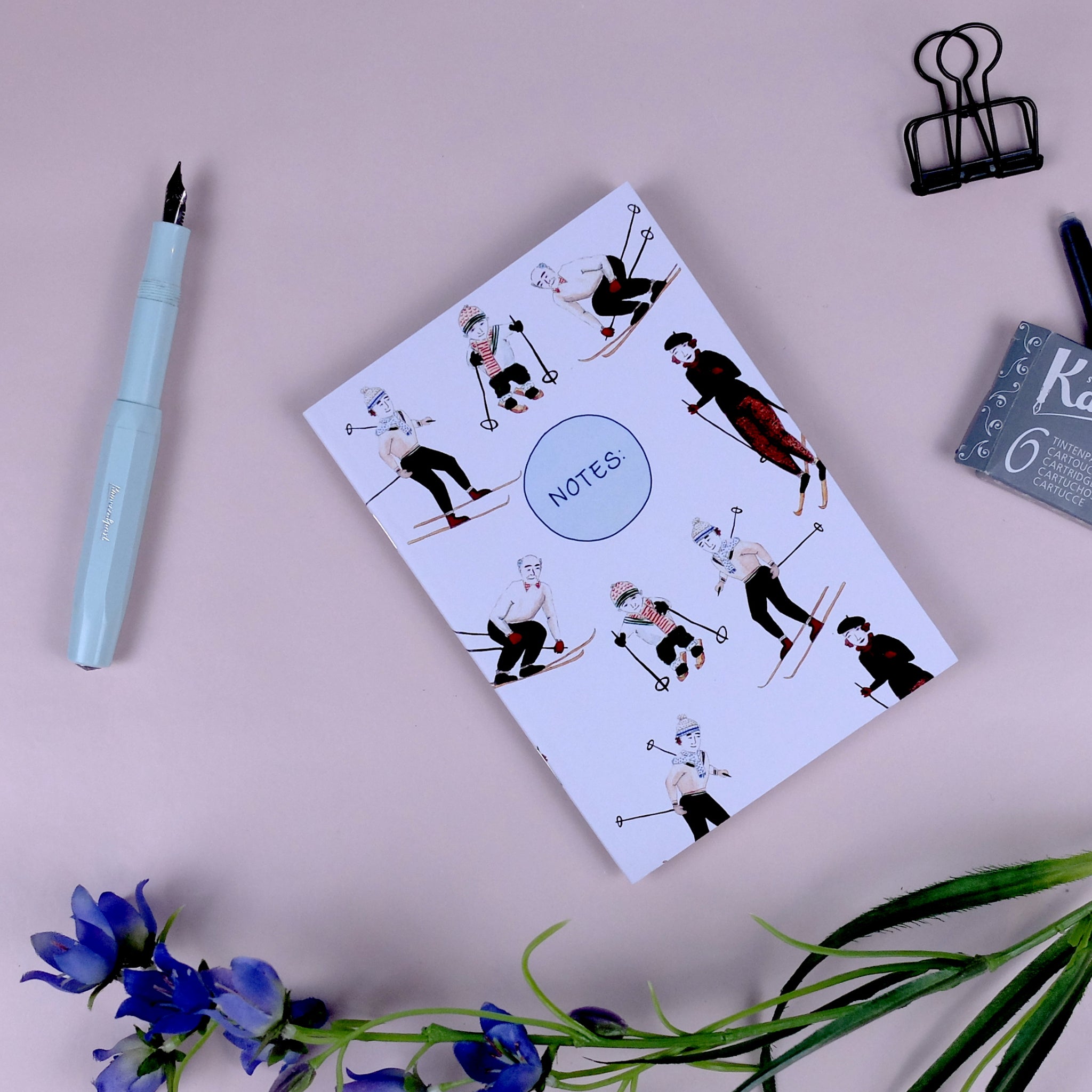 Notizbuch mit Skifahrern auf dem Cover, neben einem blauen Füller, Blumen und einer Patrone auf rosa Hintergrund.