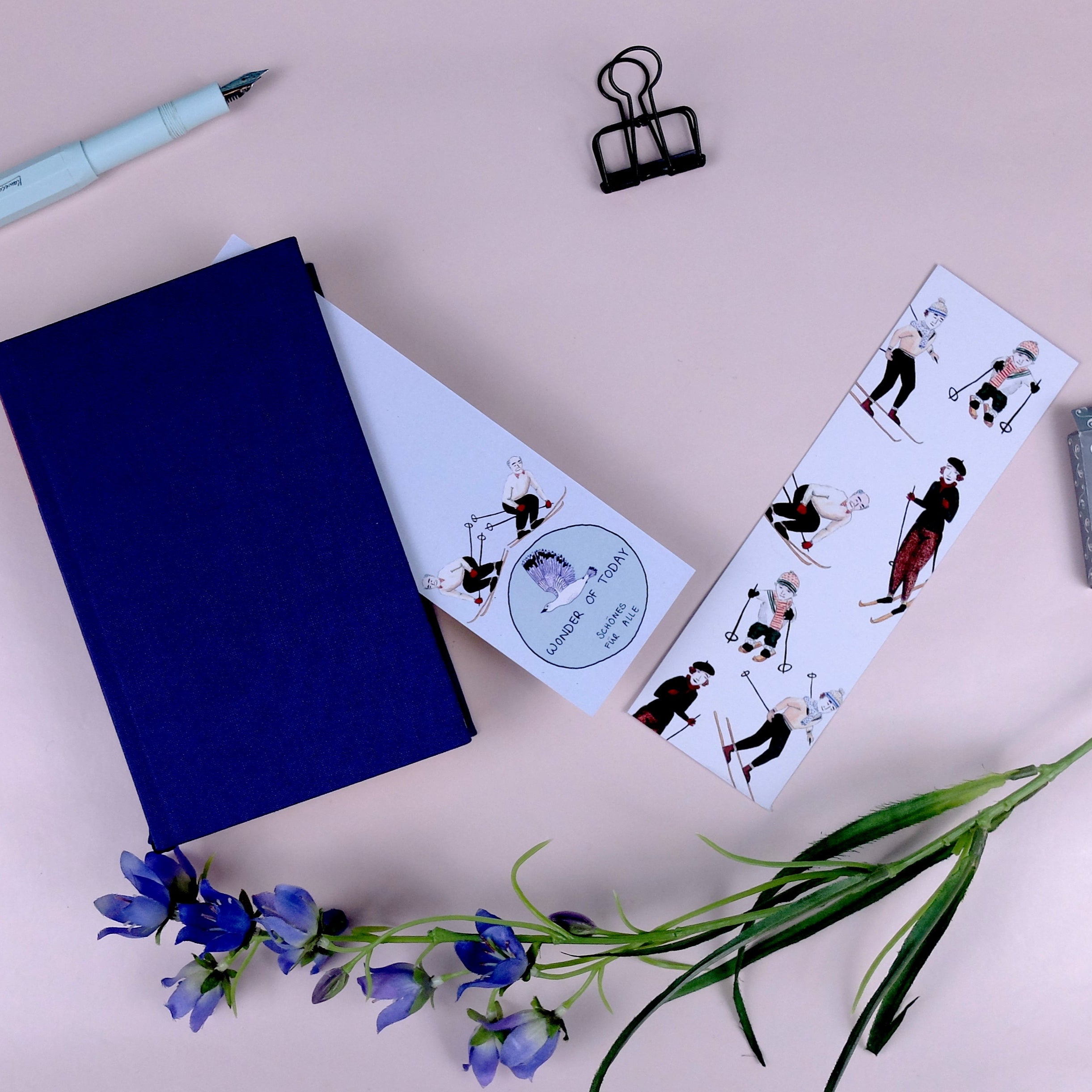 Ein blaues Notizbuch, ein Stift, eine Büroklammer, ein Radiergummi, zwei illustrierte Lesezeichen und ein Zweig mit lila Blumen auf einer hellbeigen Oberfläche.