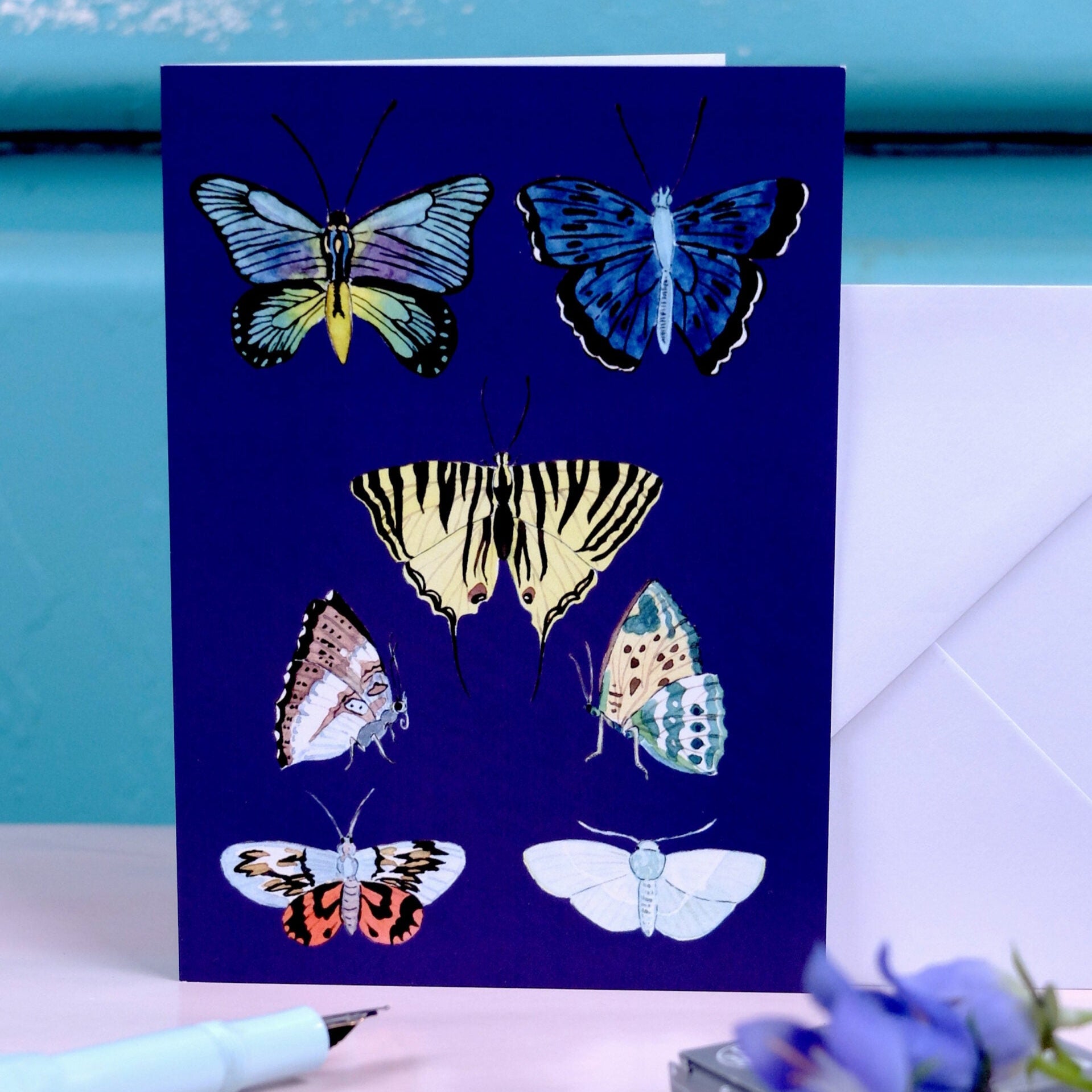 Karte mit sechs bunten Schmetterlingen auf dunkelblauem Hintergrund, daneben ein weißer Umschlag auf einem Tisch.