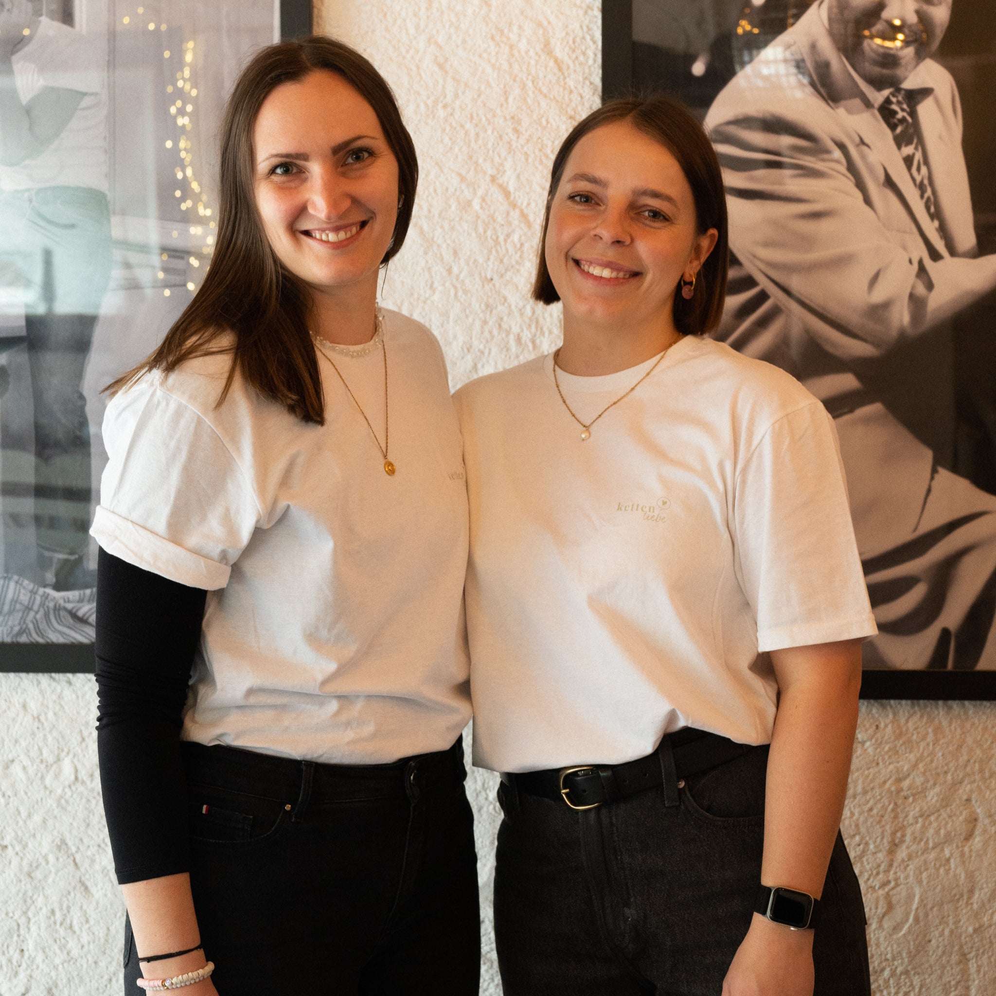 Zwei junge Frauen stehen Seite an Seite und lächeln in die Kamera. Beide tragen weiße T-Shirts und dunkle Hosen, dazu einfache Halsketten. Hinter ihnen hängt ein Schwarz-Weiß-Foto an der Wand.