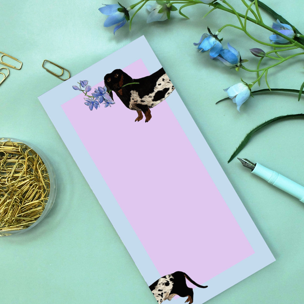 Notizblock mit lila Hintergrund und Hundeillustration, daneben eine Feder, Büroklammern und blaue Blumen.