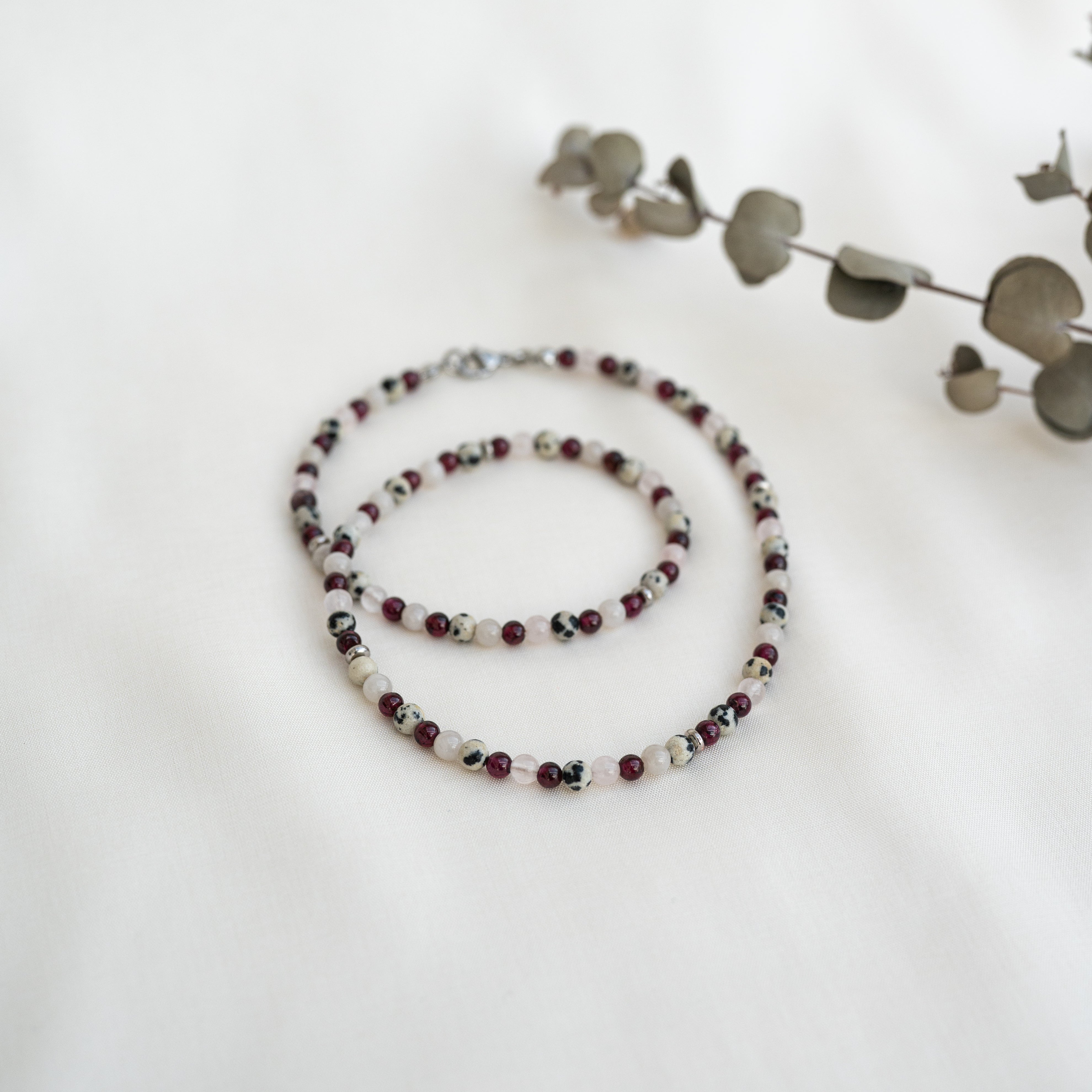 Perlenhalskette mit roten, schwarzen und weißen Steinen auf einer weißen Oberfläche, mit einem getrockneten Eukalyptuszweig im Hintergrund.