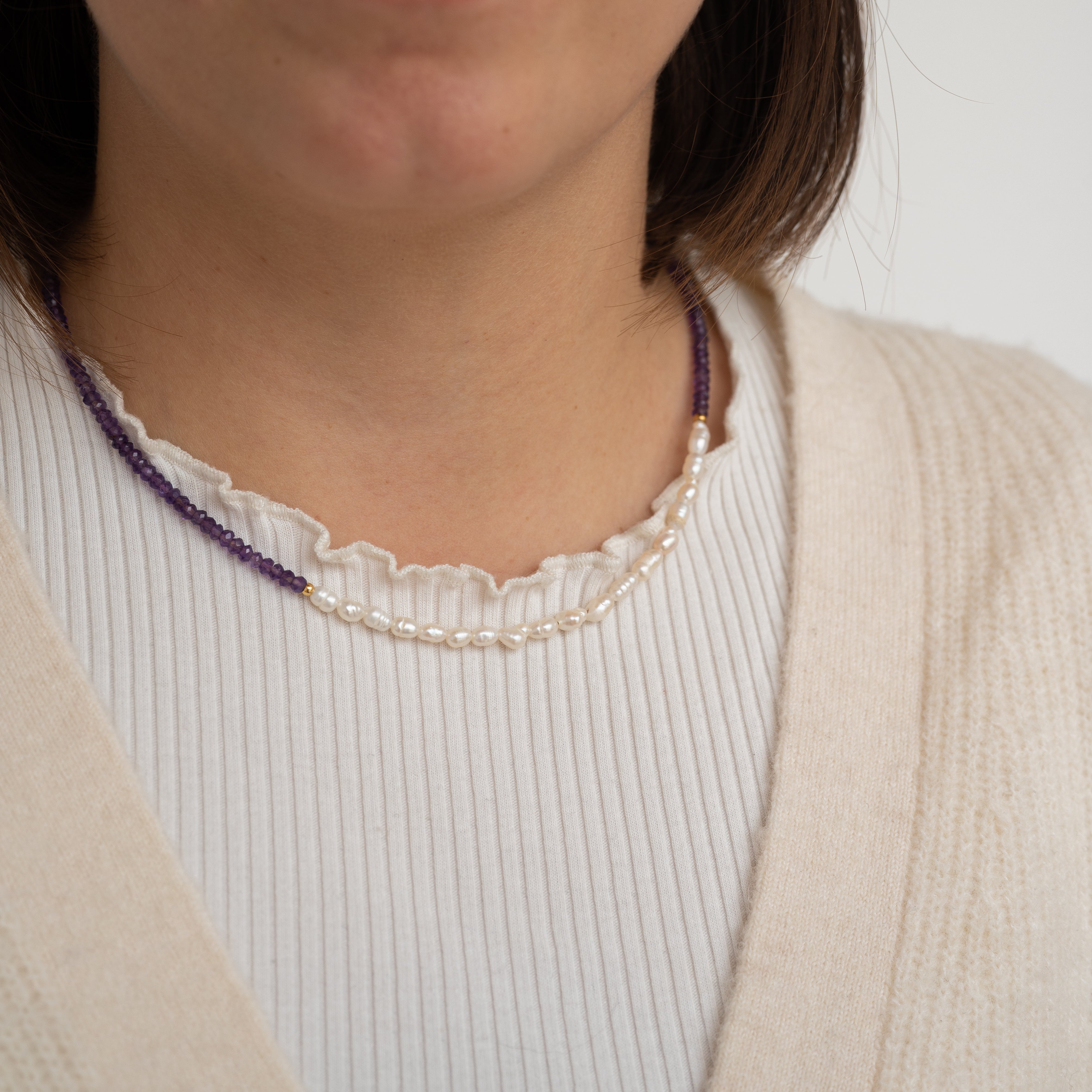Eine Frau, die einen cremefarbenen Pullover über einem weißen gerippten Oberteil trägt, mit zwei Halsketten: eine aus kleinen lila Perlen und eine aus weißen Perlen, die um ihren Hals geschichtet sind.