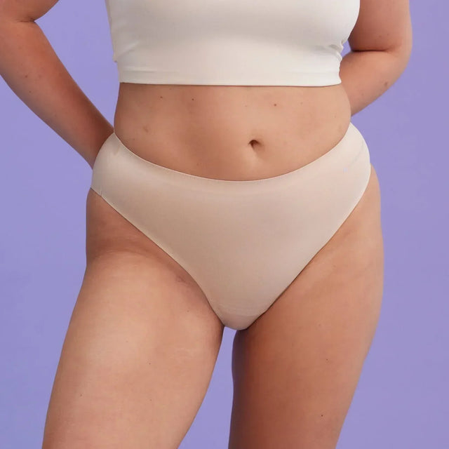 Eine Frau, die eine beigefarbene Unterwäsche mit hoher Taille und ein weißes Crop-Top trägt, steht vor einem schlichten lavendelfarbenen Hintergrund und hat die Hände auf dem Rücken.