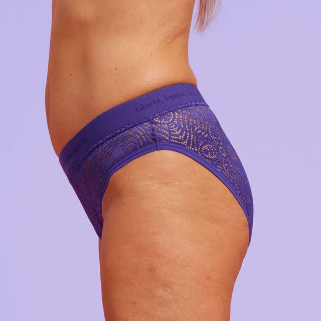 Eine Person, die ein passendes Set lila Spitzenunterwäsche trägt, wird von der Seite vor einem hellvioletten Hintergrund gezeigt, der die natürliche Hauttextur und die Kurven hervorhebt.
