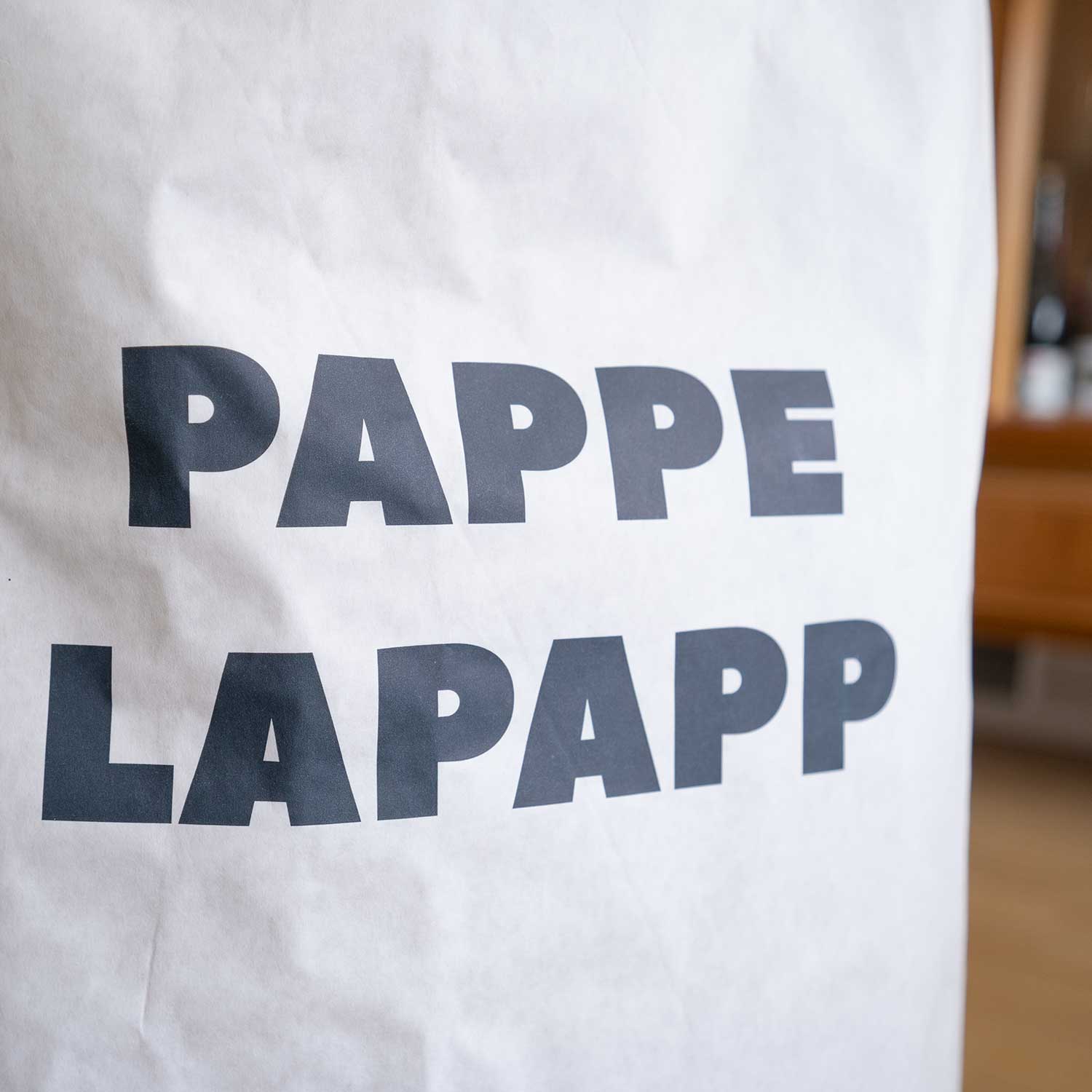 Eine weiße Papiertüte mit dem fetten schwarzen Text PAPPE LAPAPP darauf. Der Hintergrund ist unscharf, wobei etwas Braun und Weiß zu sehen sind.
