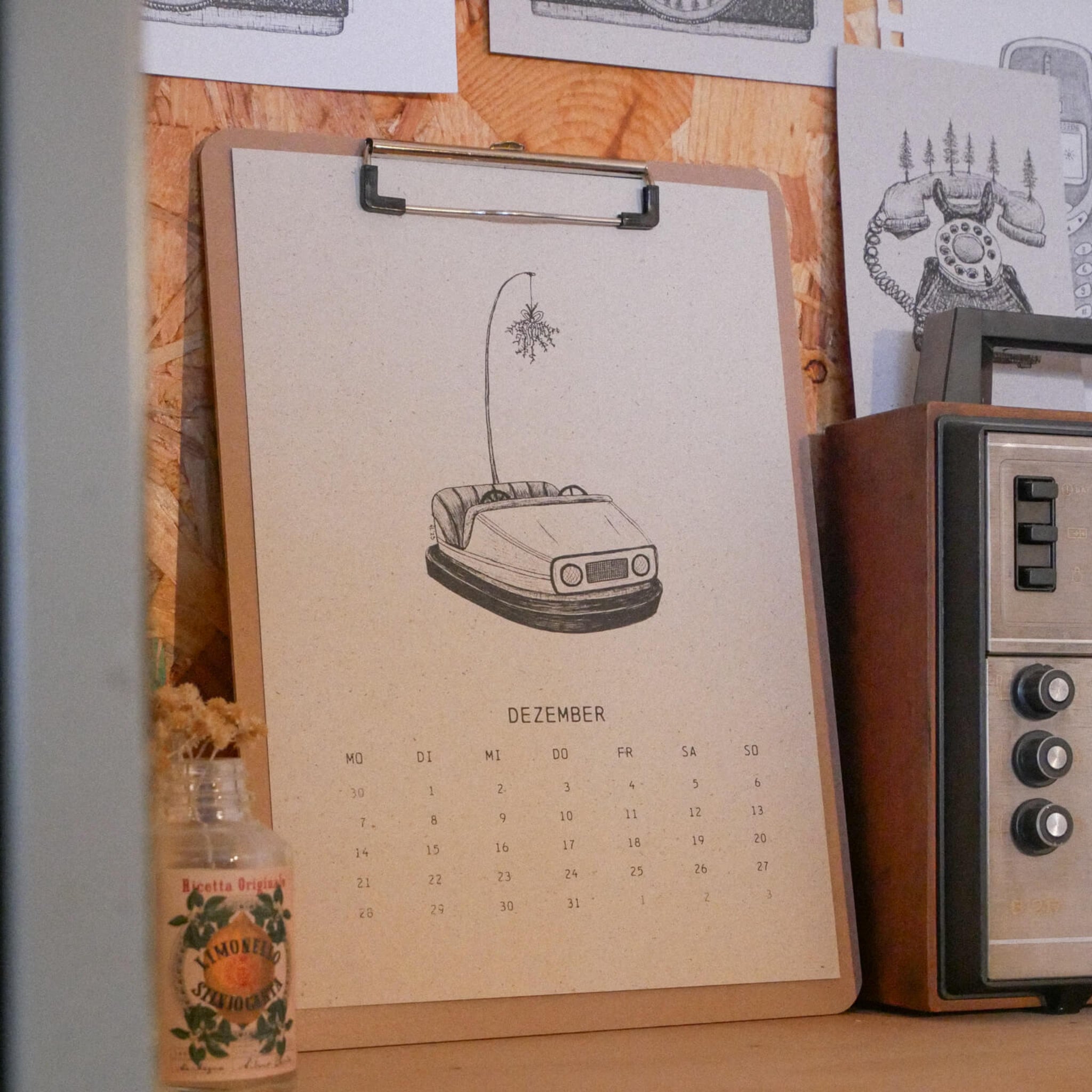 Ein Klemmbrett mit einem Dezember-Kalenderblatt, auf dem eine Zeichnung eines Autoscooters mit einem Funken darüber zu sehen ist, liegt auf einer hölzernen Unterlage, neben einem alten Radio und einer kleinen Glasflasche mit Trockenblumen.