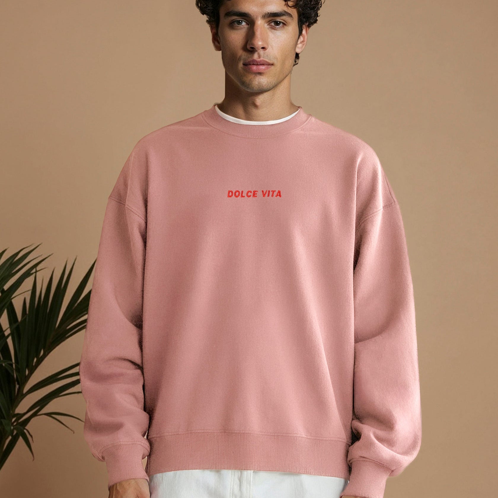 Ein junger Mann mit lockigem dunklem Haar trägt ein lockeres, rosa Sweatshirt, auf dem in kleinen roten Buchstaben DOLCE VITA steht. Er steht vor einem schlichten beigen Hintergrund mit einem Teil einer Grünpflanze auf der linken Seite.