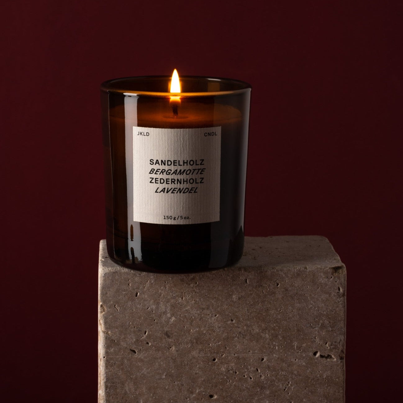 Brennende Duftkerze im Glas mit Etikett, auf dem "Sandelholz, Bergamotte, Zedernholz, Lavendel" steht, auf einem Steinblock.