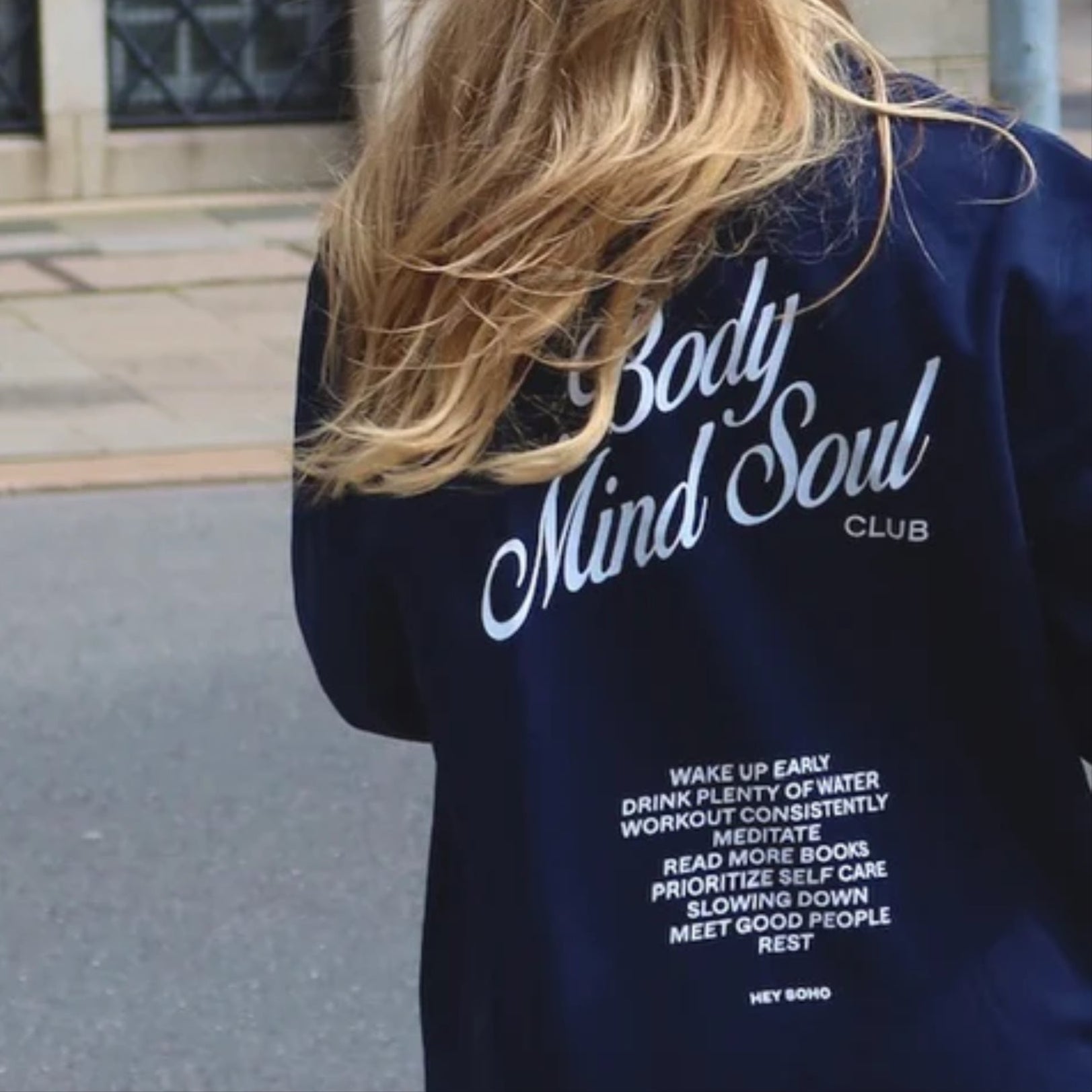 Eine Person trägt eine dunkelblaue Jacke mit der Aufschrift "Body Mind Soul Club" und einer Liste von Wellness-Tipps.