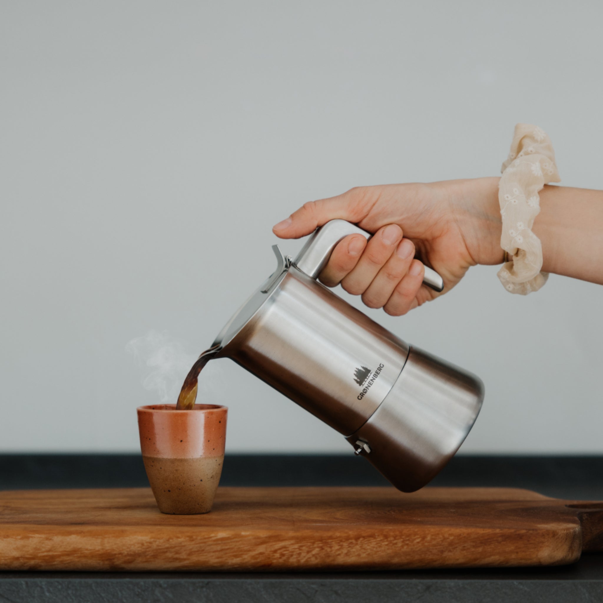 Hand pourt Kaffee aus einer silbernen Kanne in eine dampfende Tasse auf einem Holzbrett.