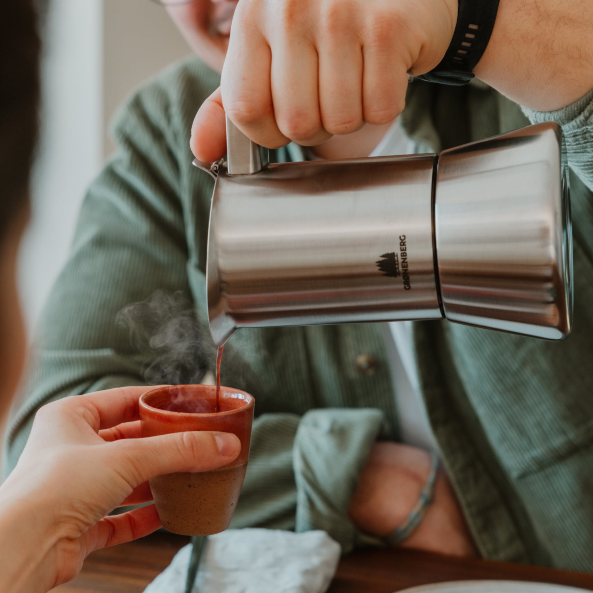 Eine Person gießt heißen Kaffee aus einer silbernen Kanne in eine kleine braune Tasse, während Dampf aufsteigt.