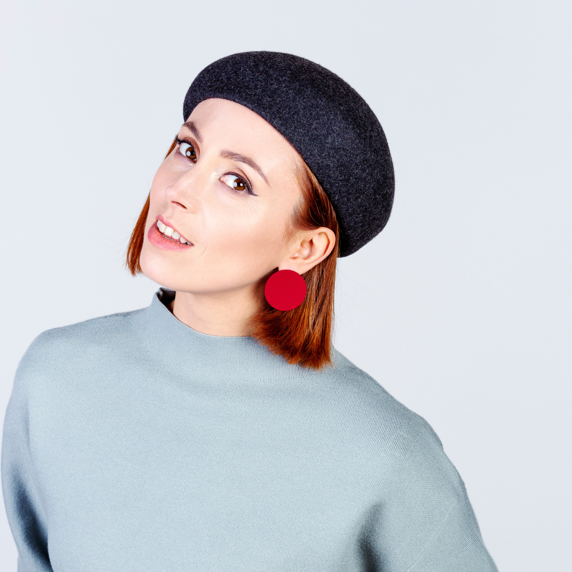 Frau mit glattem kastanienbraunem Haar, die eine dunkelgraue Baskenmütze, einen hellblauen Pullover und große rote runde Ohrringe trägt. Sie neigt ihren Kopf und lächelt leicht vor einem schlichten hellen Hintergrund.