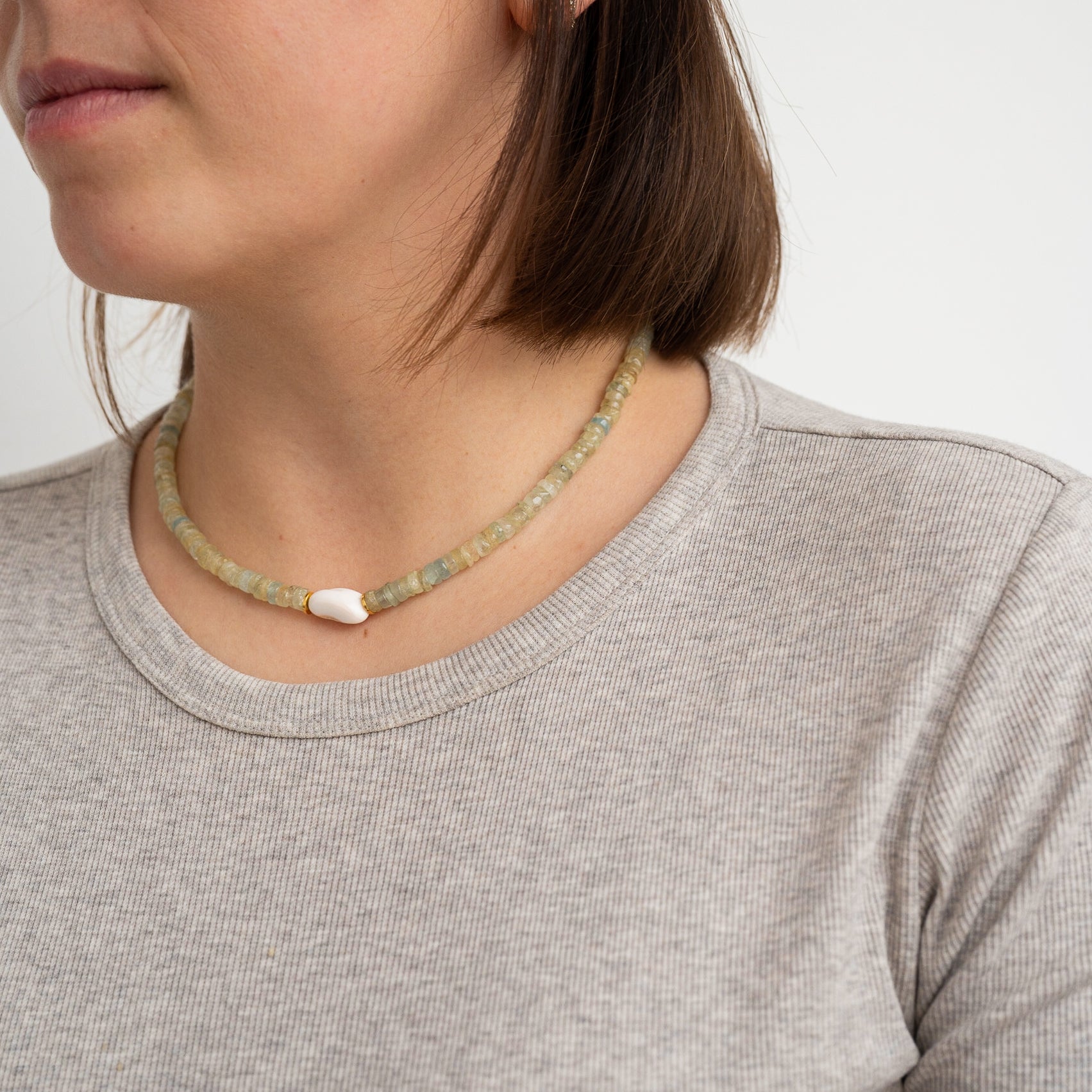 Eine Frau, die ein hellgraues geripptes Hemd und eine grüne Perlenkette mit einer einzelnen weißen Akzentperle trägt, wird von den Schultern aufwärts vor einem schlichten weißen Hintergrund gezeigt.