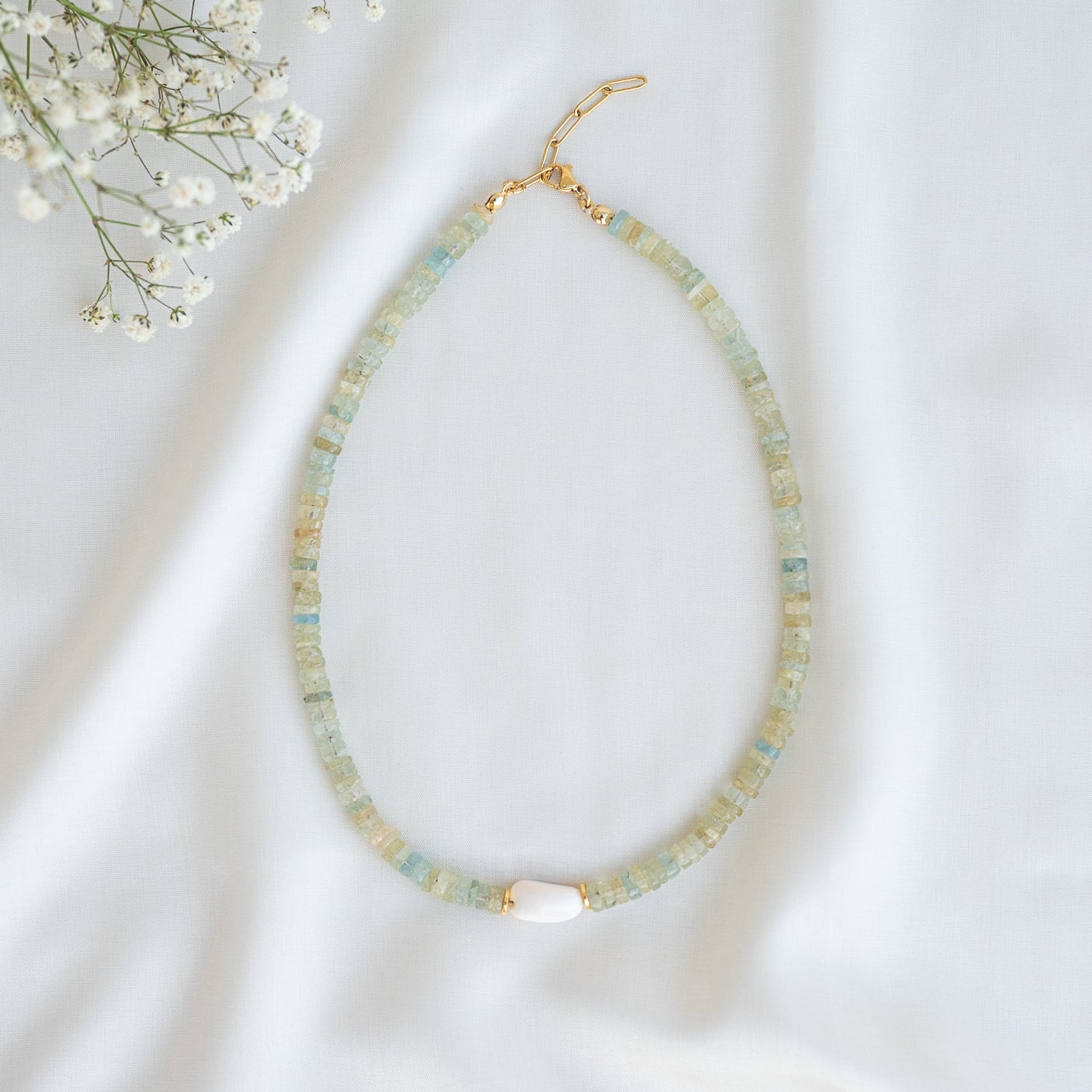 Eine Perlenkette mit hellblauen und goldfarbenen zylindrischen Perlen, mit einer einzelnen weißen Perle in der Mitte, liegt flach auf einem weißen Stoffhintergrund mit kleinen weißen Blumen in der oberen linken Ecke.