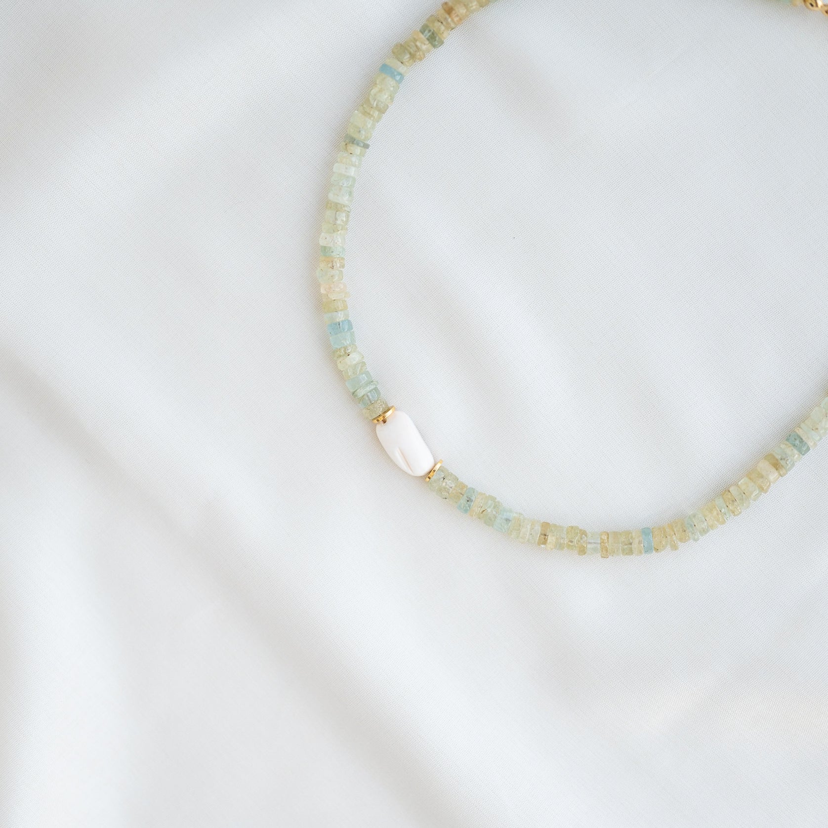 Eine zarte Perlenkette mit blassblauen und hellbeigen quadratischen Perlen, mit einer einzelnen weißen ovalen Akzentperle, die auf einem weichen weißen Stoffhintergrund präsentiert wird.