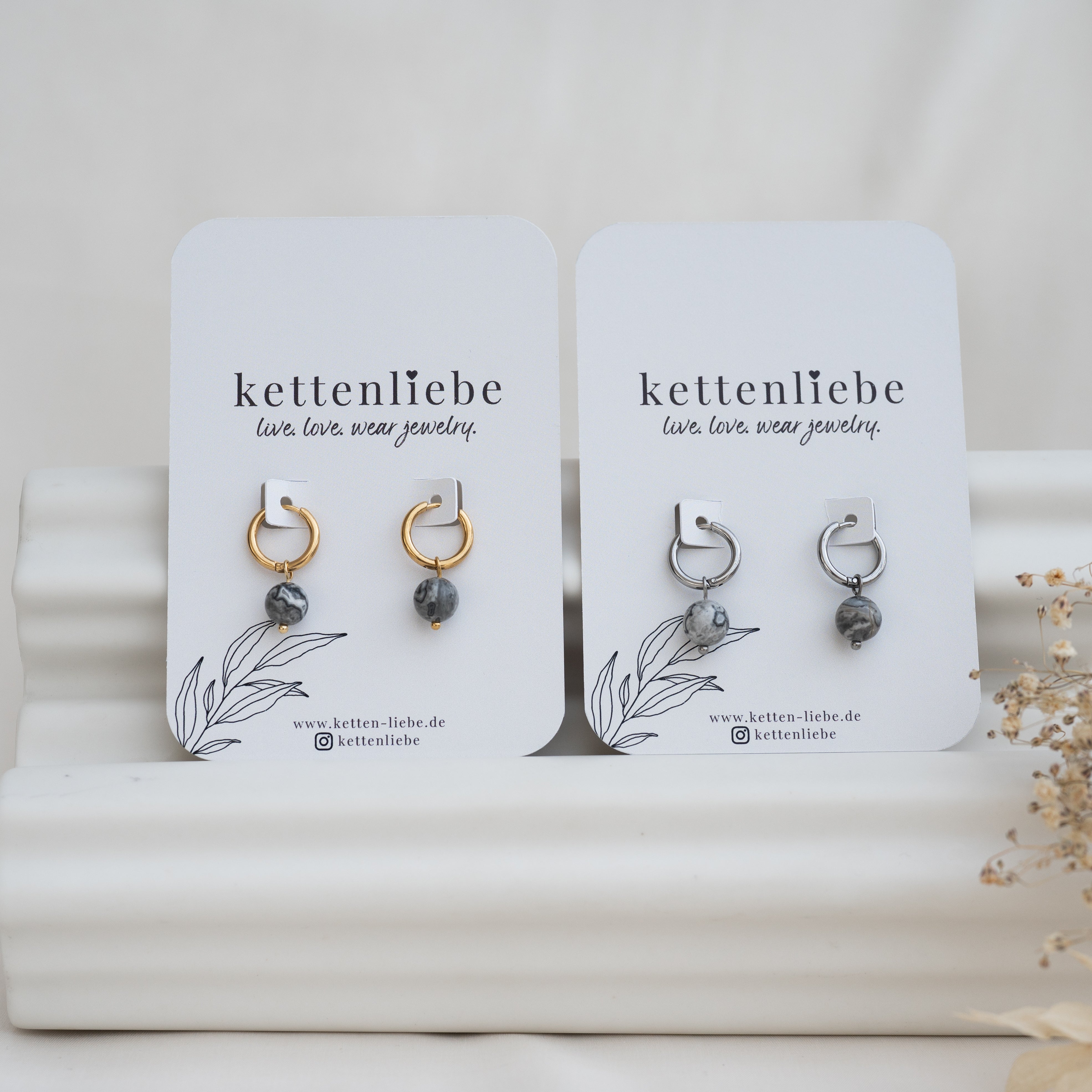 Zwei Paare von Ohrringen auf weißen Karten mit der Aufschrift kettenliebe. Ein Paar ist goldfarben mit grau marmorierten Perlen, das andere ist silberfarben mit passenden Perlen. Die Karten liegen auf einer weißen, gewellten Fläche mit Trockenblumen in der Nähe.