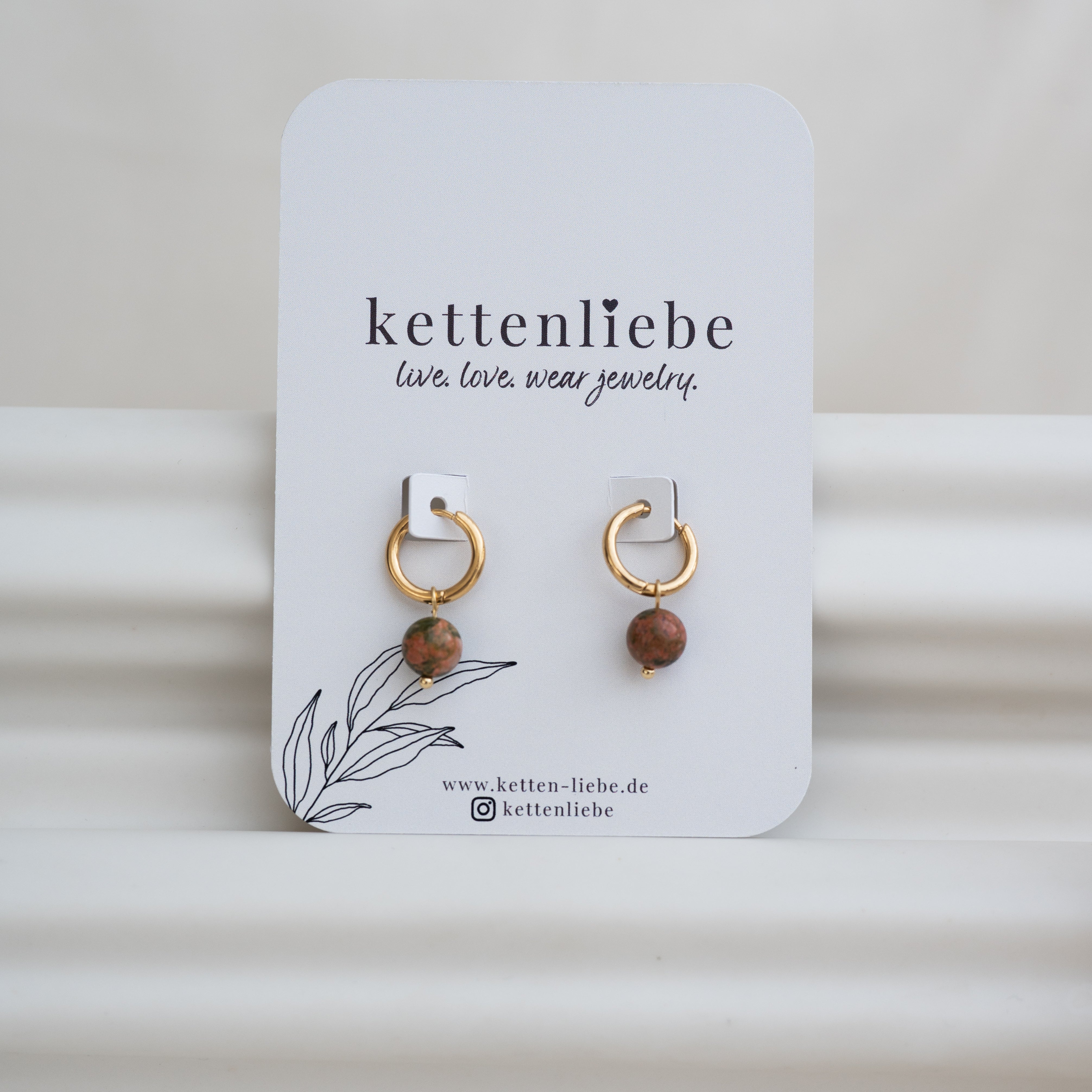 Ein Paar goldene Ohrringe mit runden, bräunlichen Steinanhängern auf einer weißen Karte mit der Aufschrift kettenliebe über einer einfachen Blattillustration und Website-Informationen.