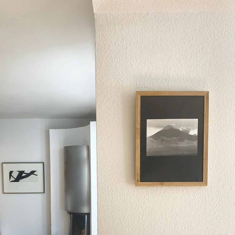 Ein gerahmtes Schwarz-Weiß-Foto eines Vulkans hängt an einer hellen Wand. Links ist eine Teilansicht eines modernen Interieurs mit einem weiteren gerahmten abstrakten Kunstwerk und einem zylindrischen Metallobjekt zu sehen.