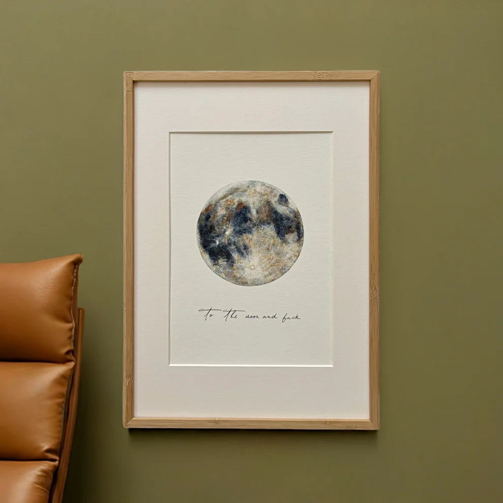 Ein gerahmtes Kunstwerk hängt an einer olivgrünen Wand über einem hellbraunen Ledersessel. Das Kunstwerk zeigt einen Mond im Aquarellstil und darunter den handgeschriebenen Satz "bis zum Mond und zurück".