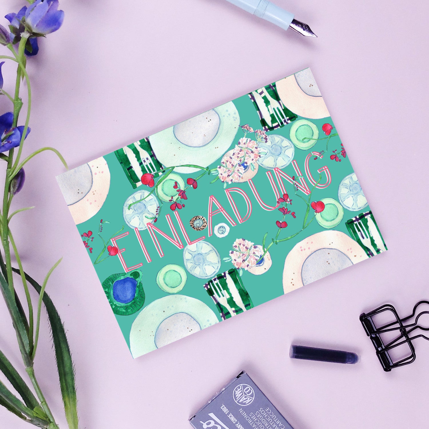 Einladungskarte mit floralen Mustern und dem Wort "EINLADUNG" auf einem Schreibtisch mit Stift und Blumen.