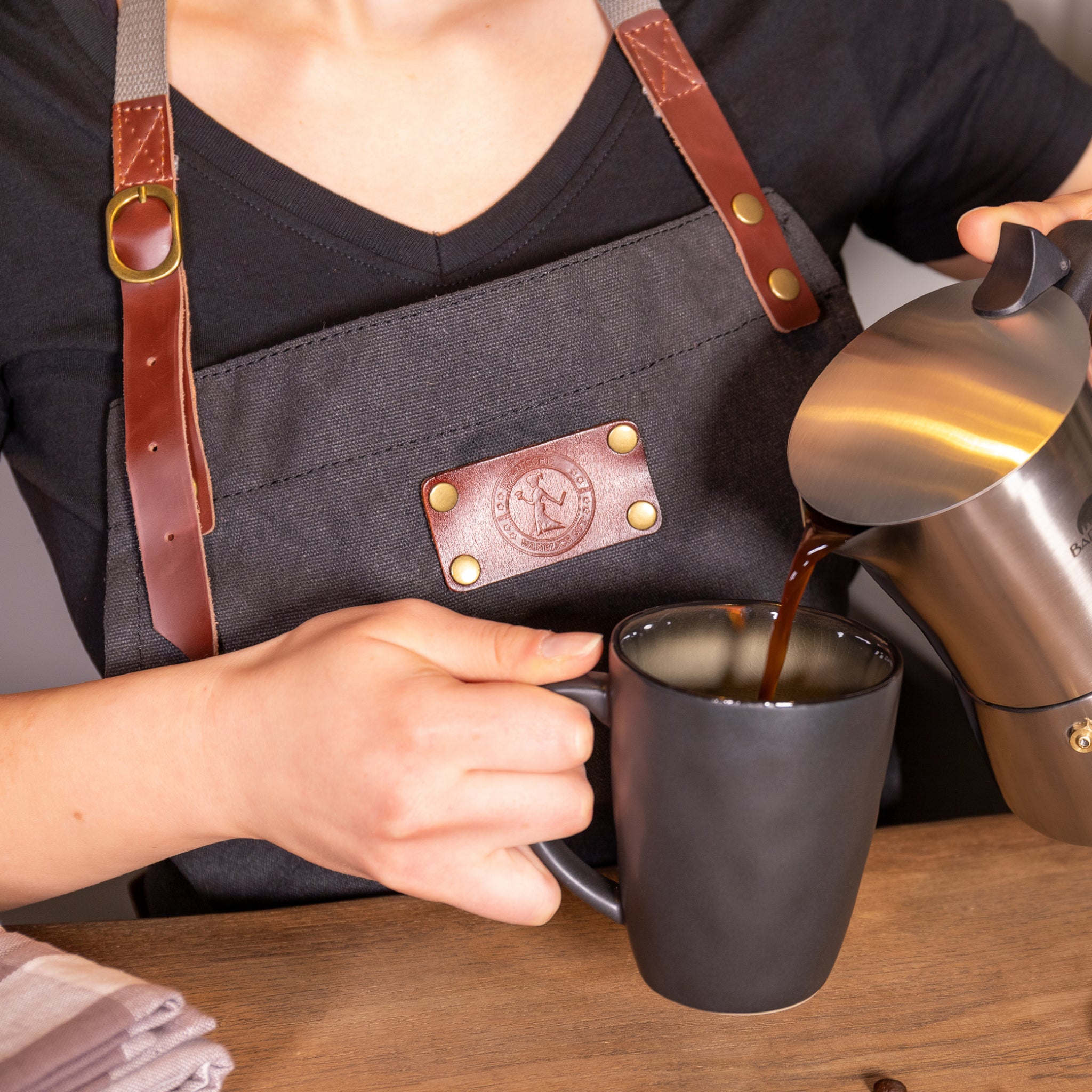 Person im schwarzen Hemd und Schürze gießt Kaffee aus einer Edelstahlkanne in eine schwarze Tasse auf einem Holztisch.