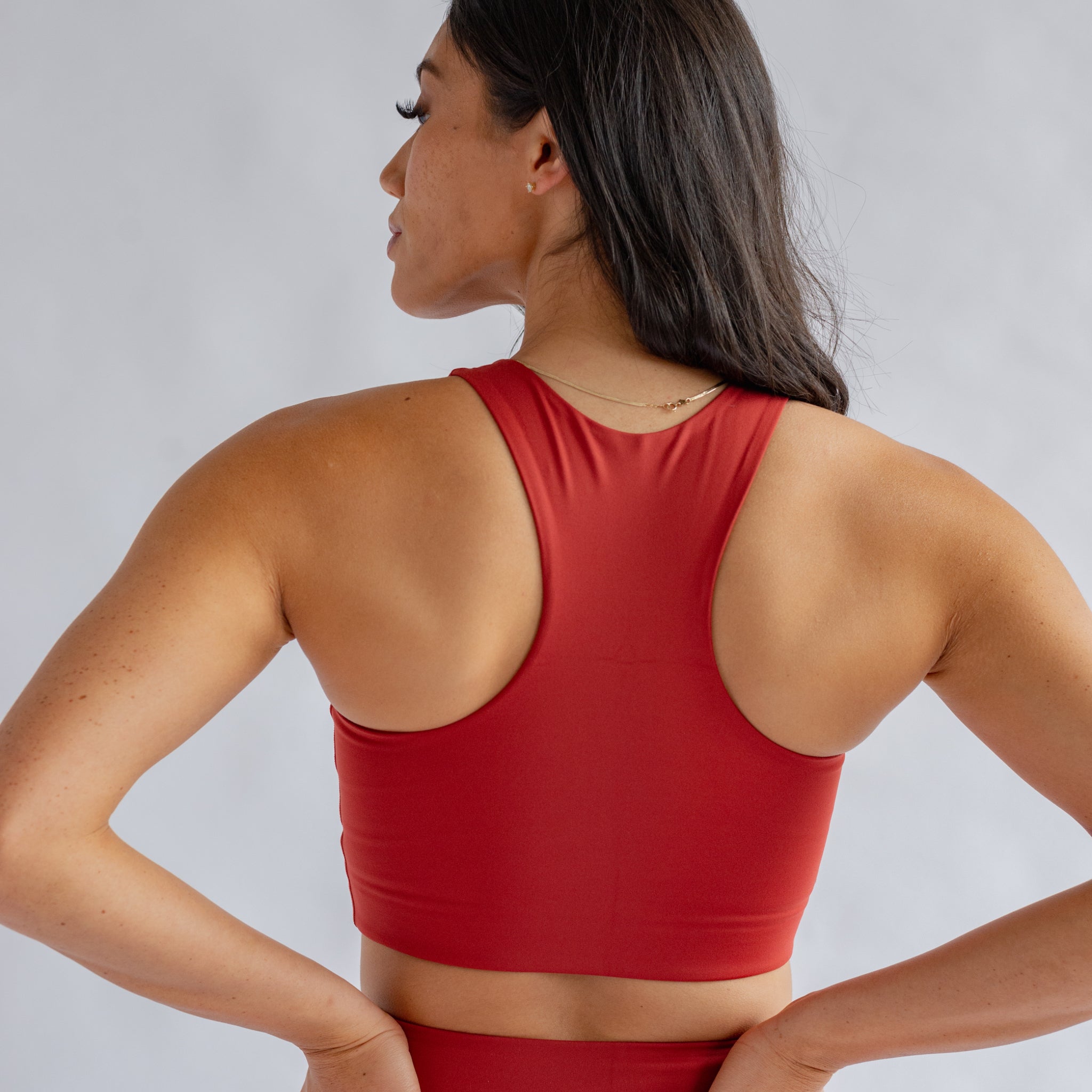 Frau von hinten in rotem Sporttop, Hände in die Hüften gestemmt, vor grauem Hintergrund.