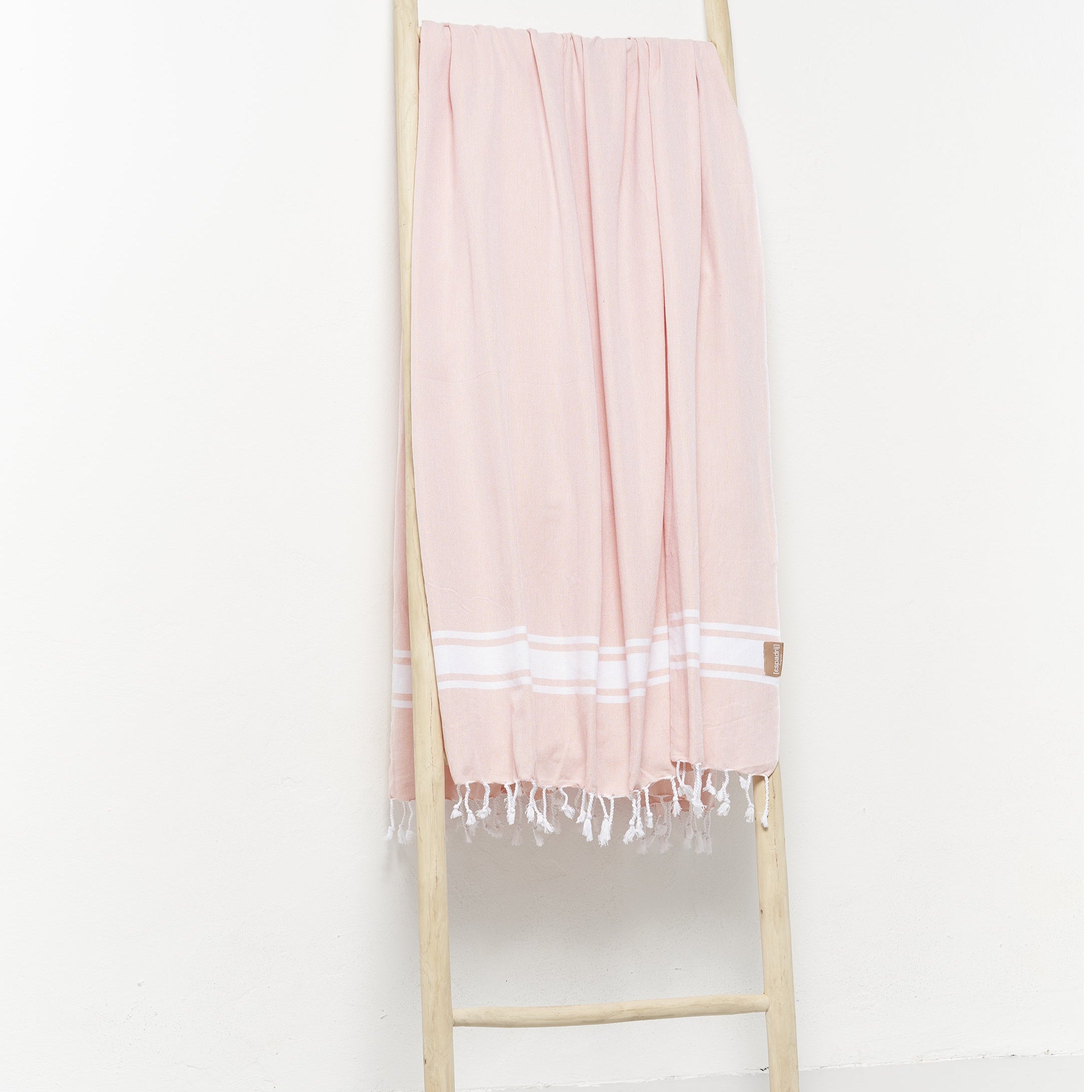 Ein pinkfarbenes Tuch mit weißen Streifen und Fransen hängt über einer Holzleiter vor einer weißen Wand.
