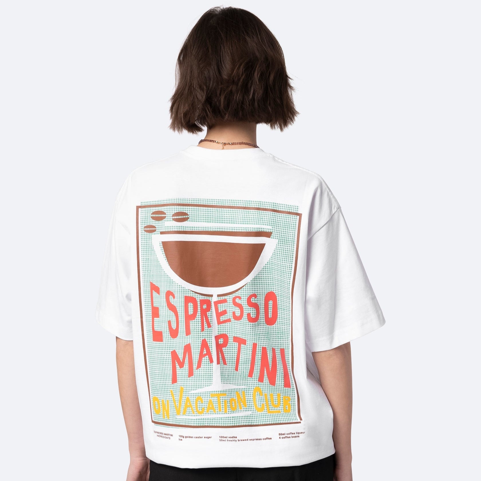 Eine Person mit kurzen braunen Haaren trägt ein weißes T-Shirt mit einer großen Grafik auf dem Rücken, auf der "Espresso Martini On Vacation Club" steht, mit einer stilisierten Martini-Glas-Illustration über dem Text.