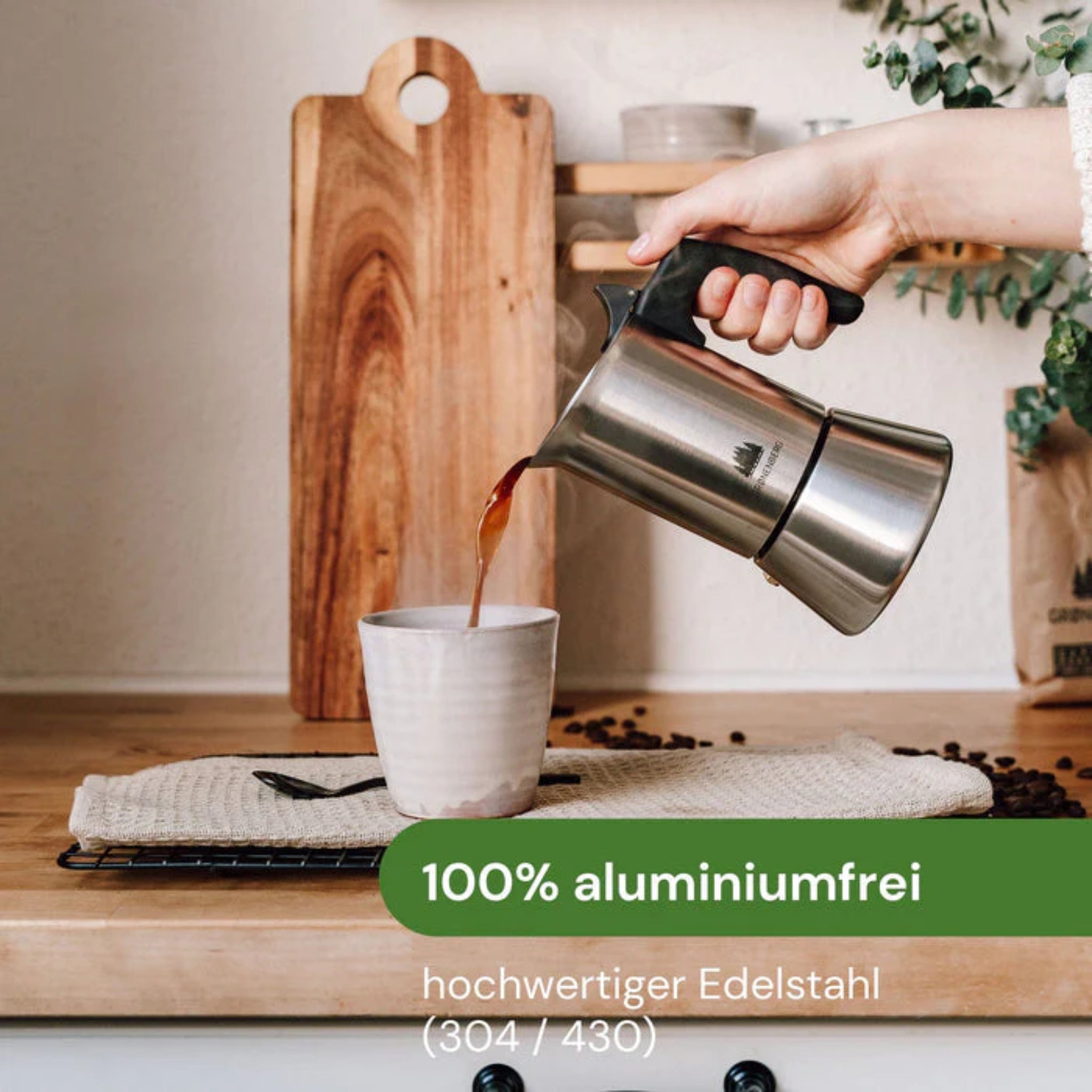 Eine Hand gießt Kaffee aus einem Edelstahlkocher in eine Tasse auf einem Holztresen. Im Hintergrund Brett und Pflanzen.