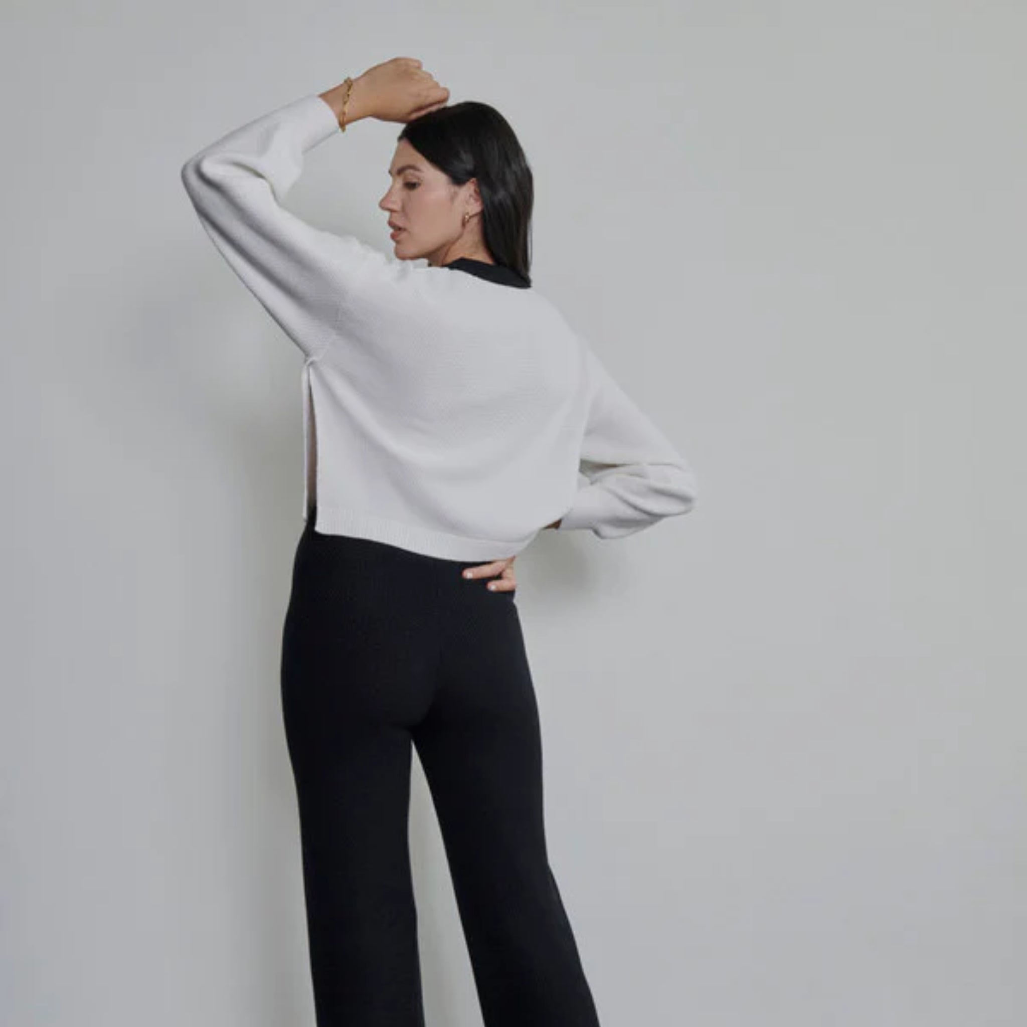 Eine Frau steht mit dem Rücken zur Kamera. Sie trägt ein weißes langärmeliges Oberteil und eine schwarze Hose mit hoher Taille. Sie hat einen Arm über dem Kopf angewinkelt und hat glattes, dunkles Haar. Der Hintergrund ist einfarbig und hell.
